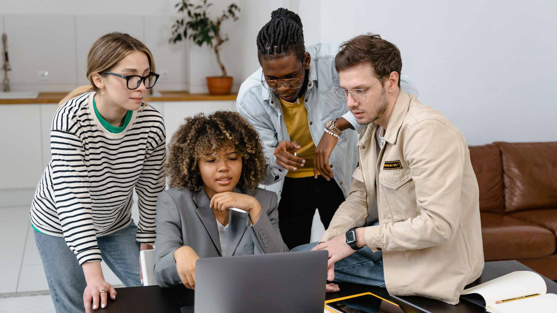 Das Bild zeigt vier Personen, die um einen Laptop herumstehen und gemeinsam an einem Projekt arbeiten. Eine Frau mit lockigem Haar sitzt und schaut konzentriert auf den Laptop, während die anderen drei, zwei Männer und eine Frau, um sie herumstehen und ebenfalls auf den Bildschirm blicken. Die Gruppe wirkt engagiert und in eine Diskussion vertieft, möglicherweise arbeiten sie an einer Präsentation oder einem kreativen Projekt. Die Umgebung ist modern und entspannt, mit einem Sofa im Hintergrund und einer schlichten Einrichtung. Die Kleidung der Personen ist lässig und spiegelt eine informelle, aber produktive Arbeitsatmosphäre wider.