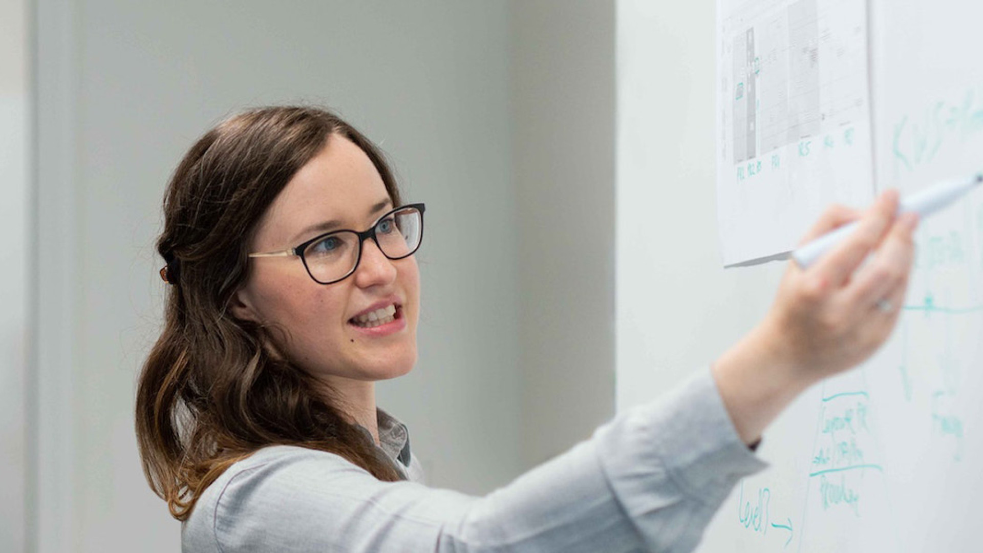 Das Bild zeigt eine Frau, die an einem Whiteboard steht und etwas darauf zeigt oder erklärt. Sie hält einen Stift in der Hand und scheint eine Präsentation oder eine Idee zu erläutern. Die Frau trägt eine Brille und ein helles Hemd. Neben ihr steht ein Mann, der aufmerksam zuhört. Er trägt ein Namensschild um den Hals und scheint in ein professionelles Gespräch mit der Frau vertieft zu sein. Die Umgebung wirkt wie ein Büro oder ein Besprechungsraum, und die beiden Personen arbeiten offenbar gemeinsam an einer Aufgabe oder einem Projekt.