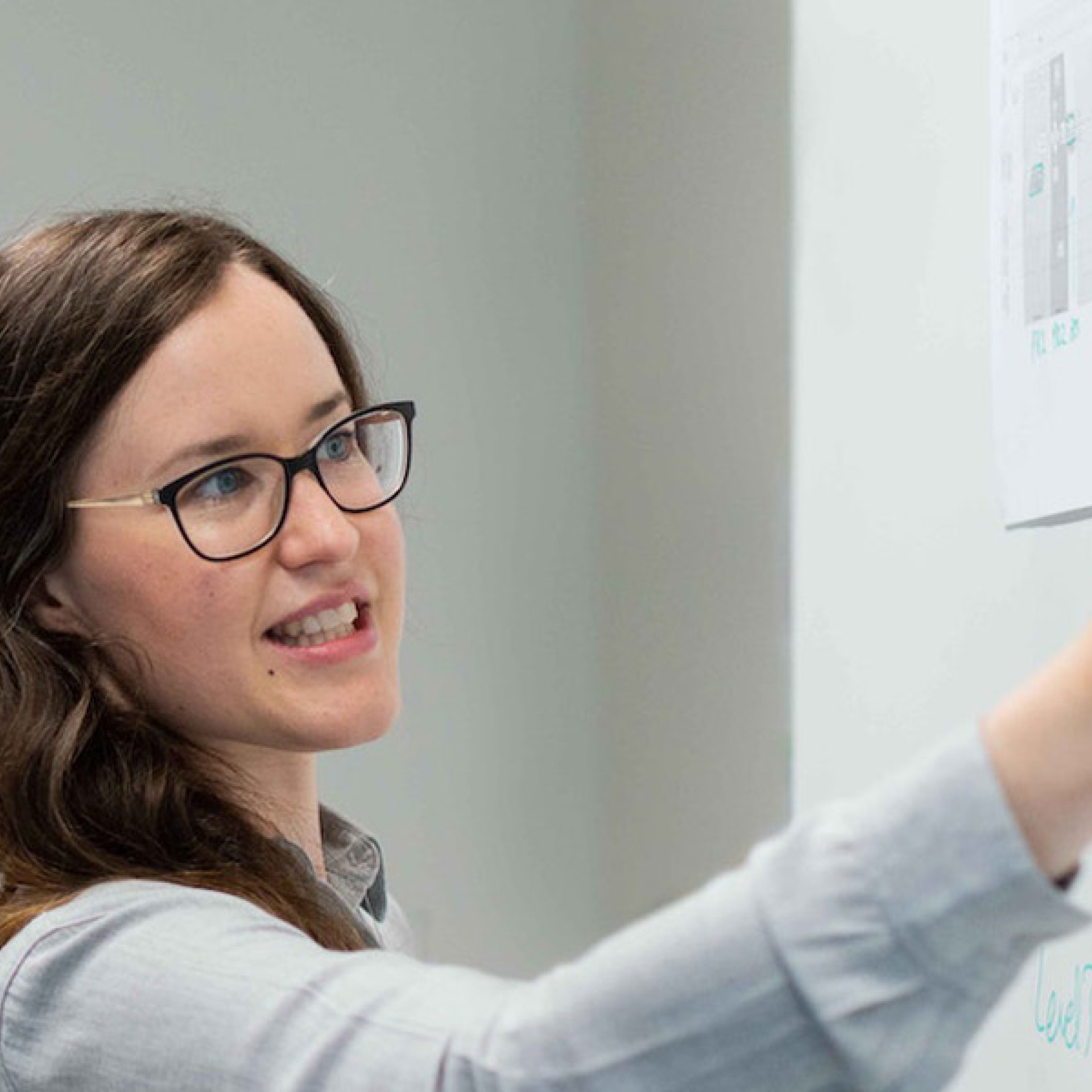 Das Bild zeigt eine Frau, die an einem Whiteboard steht und etwas darauf zeigt oder erklärt. Sie hält einen Stift in der Hand und scheint eine Präsentation oder eine Idee zu erläutern. Die Frau trägt eine Brille und ein helles Hemd. Neben ihr steht ein Mann, der aufmerksam zuhört. Er trägt ein Namensschild um den Hals und scheint in ein professionelles Gespräch mit der Frau vertieft zu sein. Die Umgebung wirkt wie ein Büro oder ein Besprechungsraum, und die beiden Personen arbeiten offenbar gemeinsam an einer Aufgabe oder einem Projekt.