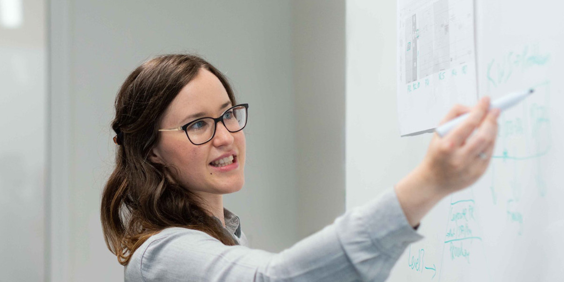 Das Bild zeigt eine Frau, die an einem Whiteboard steht und etwas darauf zeigt oder erklärt. Sie hält einen Stift in der Hand und scheint eine Präsentation oder eine Idee zu erläutern. Die Frau trägt eine Brille und ein helles Hemd. Neben ihr steht ein Mann, der aufmerksam zuhört. Er trägt ein Namensschild um den Hals und scheint in ein professionelles Gespräch mit der Frau vertieft zu sein. Die Umgebung wirkt wie ein Büro oder ein Besprechungsraum, und die beiden Personen arbeiten offenbar gemeinsam an einer Aufgabe oder einem Projekt.