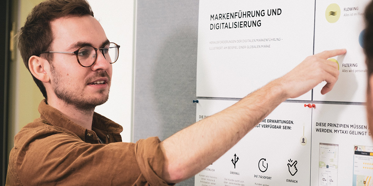 Ein junger Mann mit Brille erklärt an einem Whiteboard ein Konzept zur Markenführung und Digitalisierung. Er zeigt auf verschiedene Diagramme und Textelemente, die die Herausforderungen der digitalen Markenführung illustrieren. Die Situation vermittelt eine professionelle Lernumgebung, wie sie an der Brand University für praxisorientierte Studieninhalte typisch ist.