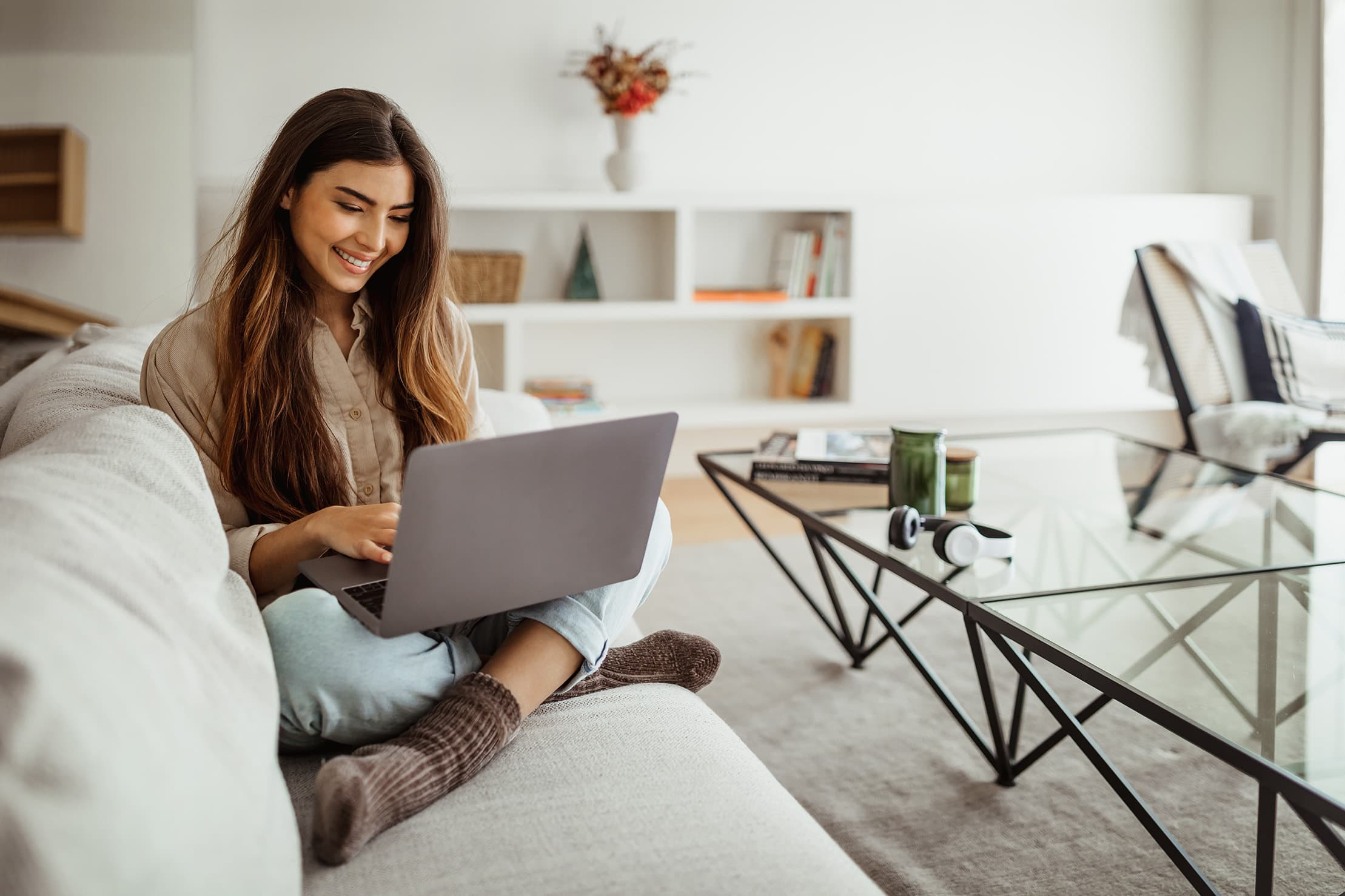 Eine junge Frau sitzt gemütlich auf einem Sofa in einem modernen, hellen Wohnzimmer und arbeitet lächelnd an ihrem Laptop. Sie trägt warme Socken und ist in eine entspannte Atmosphäre gehüllt. Diese Szene spiegelt die Flexibilität und den Komfort des ortsunabhängigen Lernens wider, das Studierenden der Brand University ermöglicht wird.