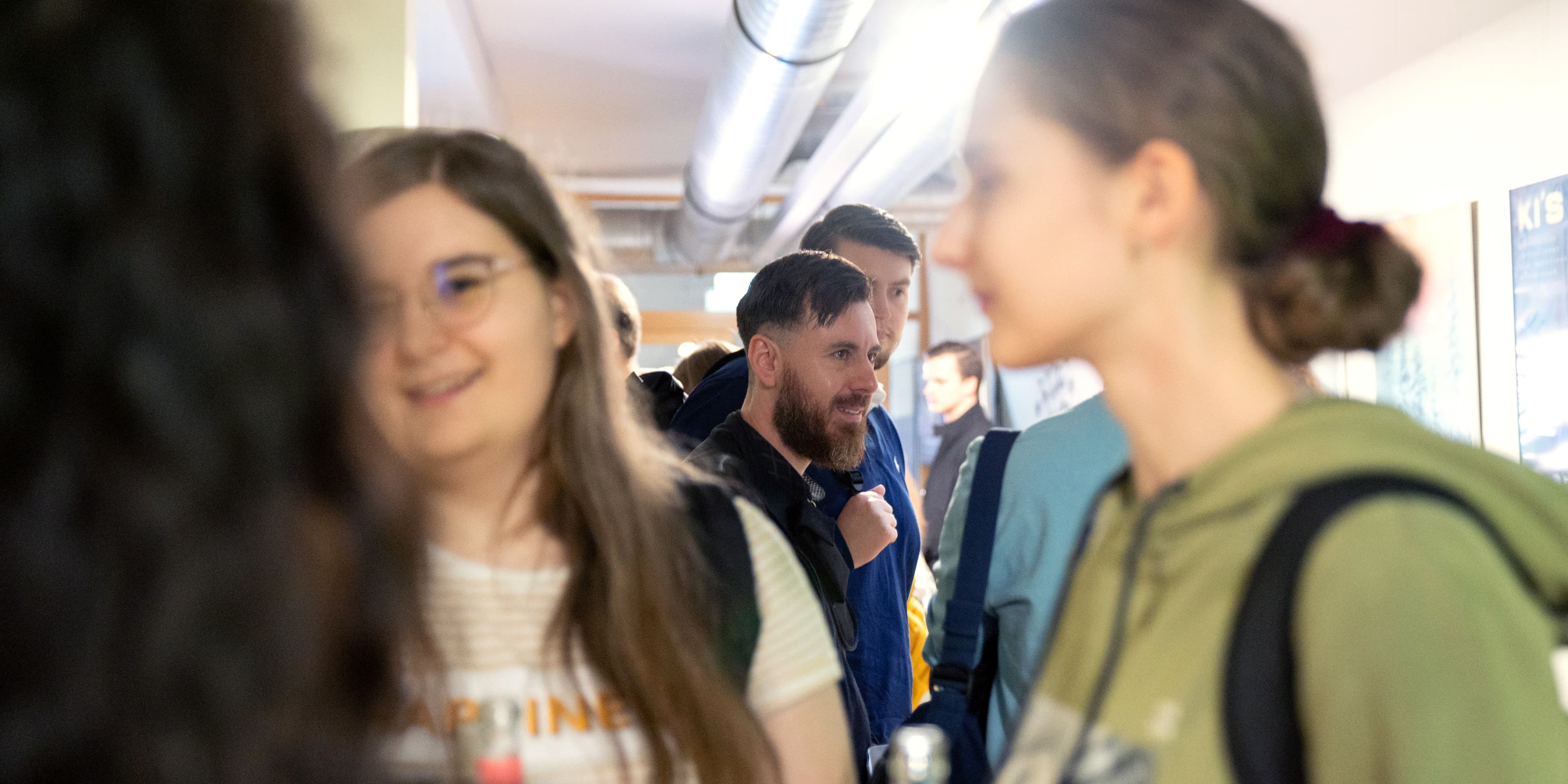 Das Bild zeigt eine Gruppe von Menschen, die in einem Flur stehen und sich unterhalten. Im Vordergrund sind zwei Frauen zu sehen, eine trägt ein weißes T-Shirt und eine Brille, die andere hat ihre Haare zu einem Dutt gebunden und trägt einen grünen Pullover. Beide halten Getränke in der Hand. Im Hintergrund sind weitere Personen zu erkennen, darunter ein Mann mit Bart, der ebenfalls in ein Gespräch vertieft ist. Die Szenerie wirkt wie eine entspannte, informelle Zusammenkunft.