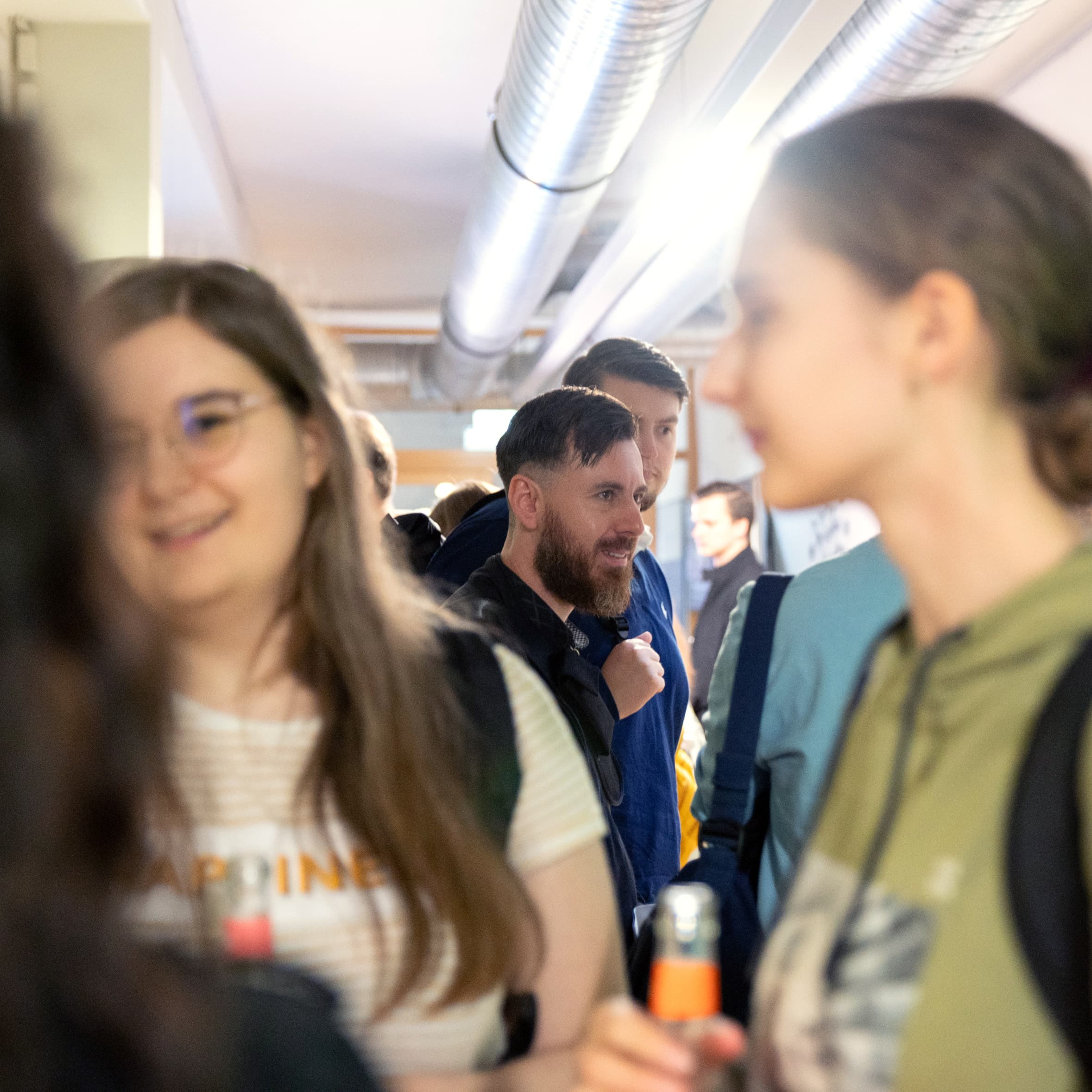 Das Bild zeigt eine Gruppe von Menschen, die in einem Flur stehen und sich unterhalten. Im Vordergrund sind zwei Frauen zu sehen, eine trägt ein weißes T-Shirt und eine Brille, die andere hat ihre Haare zu einem Dutt gebunden und trägt einen grünen Pullover. Beide halten Getränke in der Hand. Im Hintergrund sind weitere Personen zu erkennen, darunter ein Mann mit Bart, der ebenfalls in ein Gespräch vertieft ist. Die Szenerie wirkt wie eine entspannte, informelle Zusammenkunft.