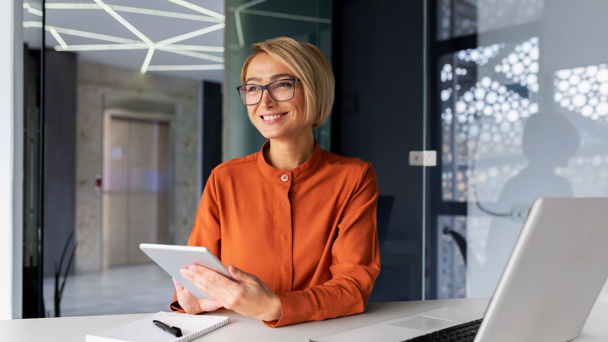 Das Bild zeigt eine Frau mit kurzen, blonden Haaren und Brille, die an einem modernen Schreibtisch sitzt. Sie trägt eine orangefarbene Bluse und hält ein Tablet in den Händen. Vor ihr liegen ein Notizblock und ein Stift auf dem Tisch, neben einem aufgeklappten Laptop. Die Umgebung wirkt modern und stilvoll, mit Glaswänden und geometrischen Deckenleuchten im Hintergrund. Die Frau lächelt freundlich und scheint in Gedanken vertieft oder in ein Gespräch involviert zu sein, was eine professionelle und konzentrierte Arbeitsatmosphäre vermittelt.