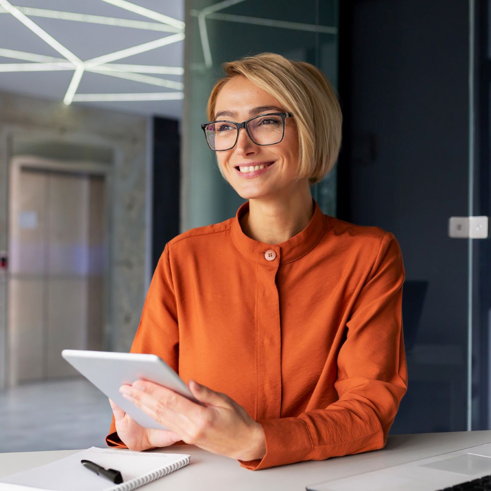 Das Bild zeigt eine Frau mit kurzen, blonden Haaren und Brille, die an einem modernen Schreibtisch sitzt. Sie trägt eine orangefarbene Bluse und hält ein Tablet in den Händen. Vor ihr liegen ein Notizblock und ein Stift auf dem Tisch, neben einem aufgeklappten Laptop. Die Umgebung wirkt modern und stilvoll, mit Glaswänden und geometrischen Deckenleuchten im Hintergrund. Die Frau lächelt freundlich und scheint in Gedanken vertieft oder in ein Gespräch involviert zu sein, was eine professionelle und konzentrierte Arbeitsatmosphäre vermittelt.