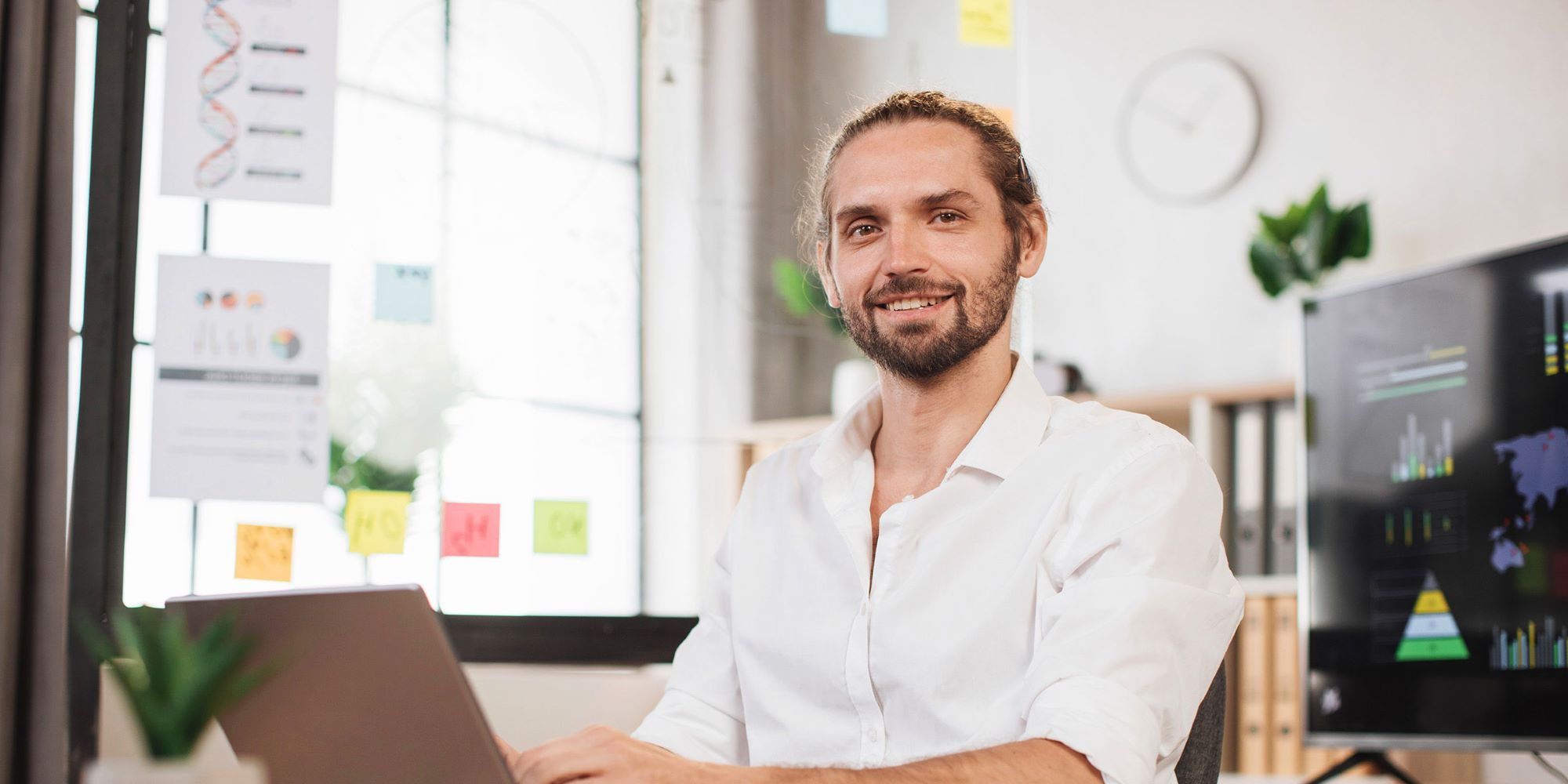 Das Bild zeigt einen Mann mit blondem Haar, das zu einem kleinen Knoten zusammengebunden ist, und einem kurzen Bart. Er sitzt an einem Schreibtisch in einem modernen Büro und lächelt freundlich in die Kamera. Der Mann trägt ein weißes Hemd mit hochgekrempelten Ärmeln und arbeitet an einem Laptop. Im Hintergrund sind Glaswände mit angehefteten Notizen und Diagrammen, eine Pflanze sowie ein großer Bildschirm zu sehen, auf dem Datenvisualisierungen angezeigt werden. Die Atmosphäre wirkt professionell und gleichzeitig kreativ, was auf ein Arbeitsumfeld hindeutet, in dem strategische Planung oder Projektarbeit im Mittelpunkt stehen.