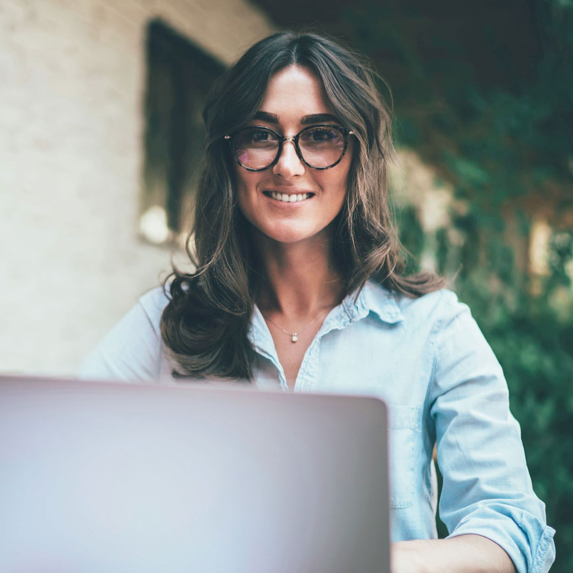 Das Bild zeigt eine Frau mit langen, welligen Haaren und Brille, die vor einem Laptop sitzt und freundlich in die Kamera lächelt. Sie trägt ein hellblaues Hemd und scheint in einer entspannten Umgebung zu arbeiten, möglicherweise im Freien oder in einem Café, da im Hintergrund Pflanzen und eine Wand zu sehen sind. Neben ihrem Laptop liegt ein Smartphone, was darauf hindeutet, dass sie möglicherweise multitasking-fähig ist oder in einer mobilen Arbeitsumgebung tätig ist. Die Atmosphäre wirkt ruhig und angenehm, während die Frau konzentriert und gleichzeitig entspannt aussieht.