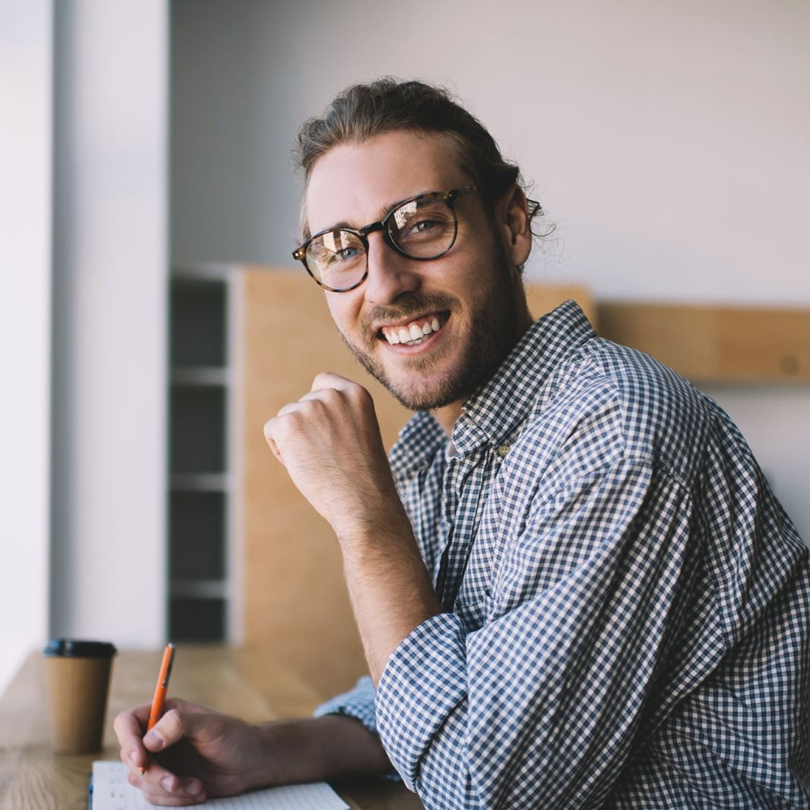 Das Bild zeigt einen jungen Mann mit Brille und zusammengebundenem Haar, der an einem Schreibtisch sitzt und freundlich in die Kamera lächelt. Er trägt ein kariertes Hemd und hält einen Stift in der Hand, während er an einem Blatt Papier schreibt. Auf dem Tisch vor ihm steht ein Becher, vermutlich mit Kaffee oder Tee. Die Umgebung wirkt schlicht und modern, mit hellen Holzmöbeln und viel natürlichem Licht, das durch ein Fenster auf der linken Seite fällt. Der Mann wirkt entspannt und konzentriert, als wäre er in seine Arbeit vertieft.