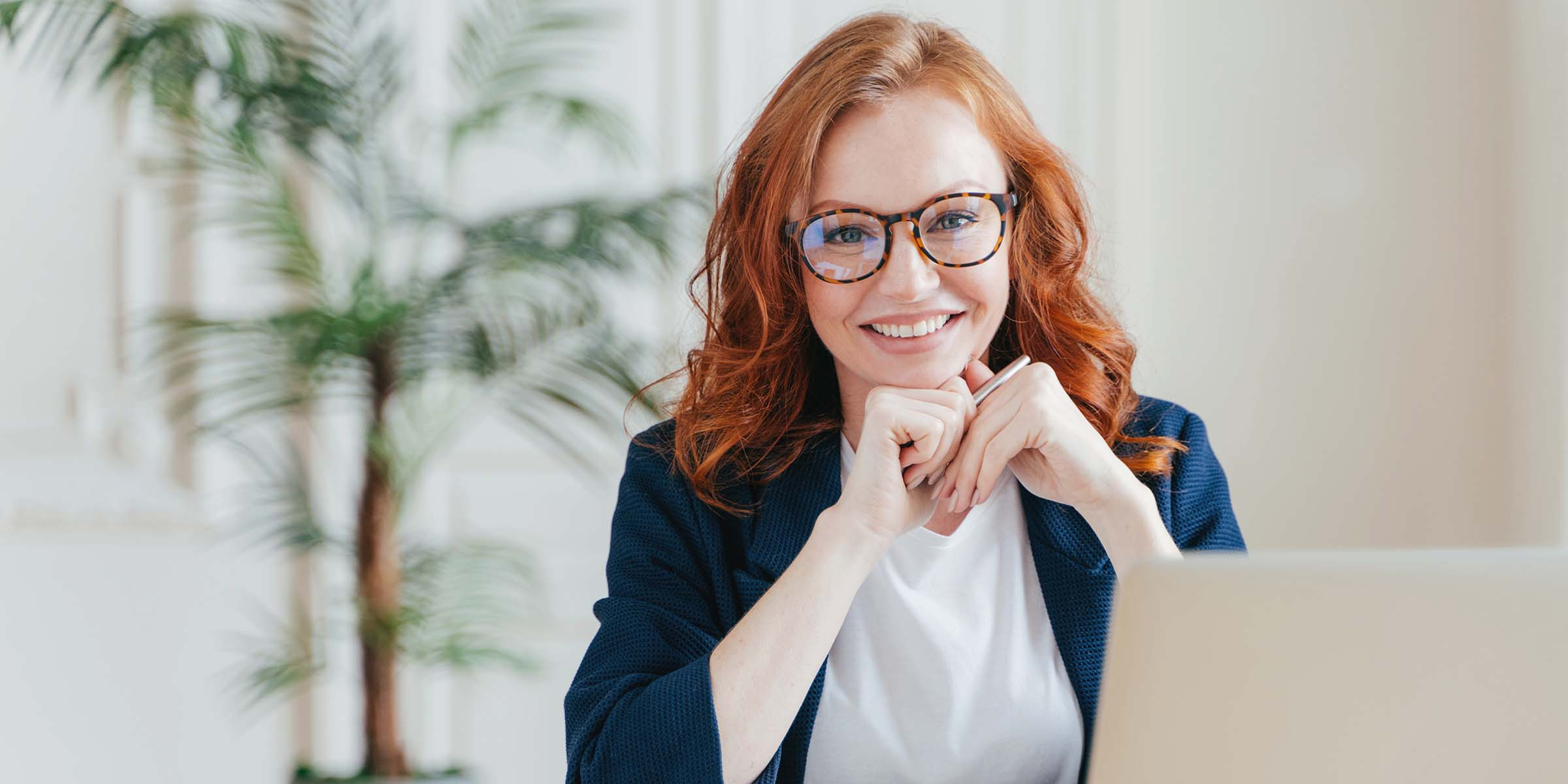 Das Bild zeigt eine junge Frau mit rotbraunen Haaren und Brille, die freundlich in die Kamera lächelt. Sie sitzt an einem Schreibtisch, stützt ihren Kopf auf ihre Hände und blickt konzentriert nach vorne. Die Frau trägt ein dunkelblaues Jackett über einem weißen Oberteil. Im Hintergrund ist eine grüne Zimmerpflanze zu sehen, was eine entspannte und freundliche Atmosphäre vermittelt. Neben ihr steht ein Laptop, was darauf hindeutet, dass sie möglicherweise an einer Arbeitssitzung teilnimmt oder sich in einer Arbeitsumgebung befindet.