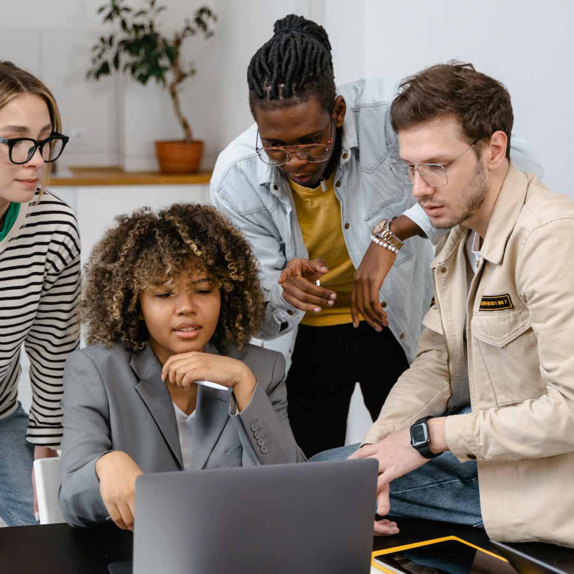 Das Bild zeigt vier Personen, die um einen Laptop herumstehen und gemeinsam an einem Projekt arbeiten. Eine Frau mit lockigem Haar sitzt und schaut konzentriert auf den Laptop, während die anderen drei, zwei Männer und eine Frau, um sie herumstehen und ebenfalls auf den Bildschirm blicken. Die Gruppe wirkt engagiert und in eine Diskussion vertieft, möglicherweise arbeiten sie an einer Präsentation oder einem kreativen Projekt. Die Umgebung ist modern und entspannt, mit einem Sofa im Hintergrund und einer schlichten Einrichtung. Die Kleidung der Personen ist lässig und spiegelt eine informelle, aber produktive Arbeitsatmosphäre wider.