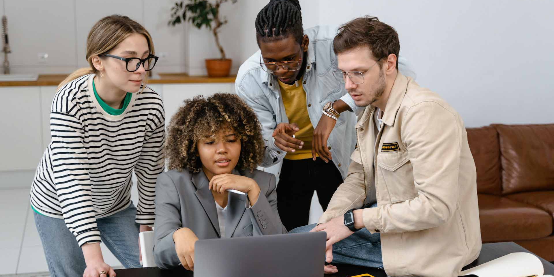 Das Bild zeigt vier Personen, die um einen Laptop herumstehen und gemeinsam an einem Projekt arbeiten. Eine Frau mit lockigem Haar sitzt und schaut konzentriert auf den Laptop, während die anderen drei, zwei Männer und eine Frau, um sie herumstehen und ebenfalls auf den Bildschirm blicken. Die Gruppe wirkt engagiert und in eine Diskussion vertieft, möglicherweise arbeiten sie an einer Präsentation oder einem kreativen Projekt. Die Umgebung ist modern und entspannt, mit einem Sofa im Hintergrund und einer schlichten Einrichtung. Die Kleidung der Personen ist lässig und spiegelt eine informelle, aber produktive Arbeitsatmosphäre wider.