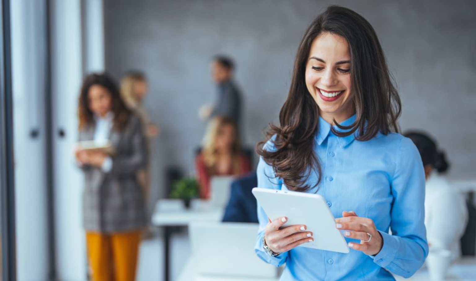 Eine Absolventin des MBA Brand Marketing – Customer Experience Management in einem modernen Büro lächelt und hält ein Tablet in den Händen. Sie trägt ein blaues Hemd und steht vor einer Gruppe von Personen, die im Hintergrund arbeiten. Die Atmosphäre ist professionell und dynamisch, mit hellen, modernen Büromöbeln und großen Fenstern.