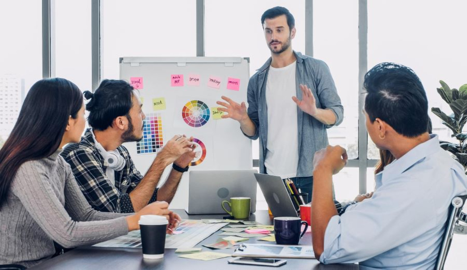 Alt-Text: Eine Gruppe von fünf Personen sitzt in einem modernen Besprechungsraum, während ein Mann vor einem Flipchart steht und eine Präsentation hält. Auf dem Flipchart sind Farbkreise und Haftnotizen zu sehen. Die anderen Personen hören aufmerksam zu und nehmen Notizen oder trinken Kaffee. Auf dem Tisch liegen Papiere, Laptops und Tassen. Das Bild vermittelt eine kreative und kollaborative Atmosphäre. Es geht um das Thema Branding.