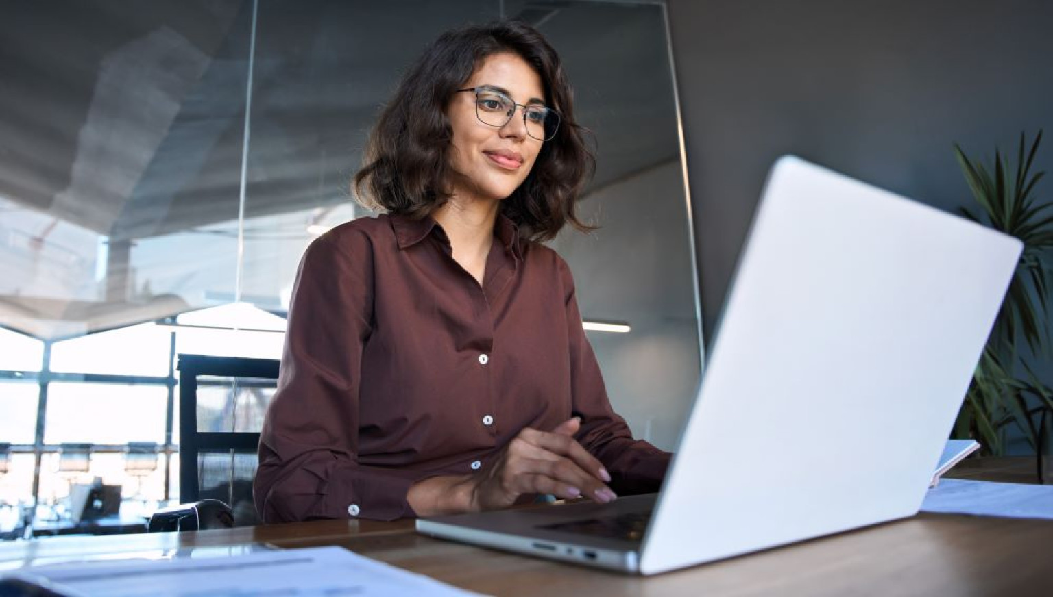 Eine Absolventin des MBA E-Commerce sitzt an einem Schreibtisch und arbeitet an ihrem Laptop. Sie trägt ein braunes Hemd und eine Brille und lächelt leicht, während sie konzentriert auf den Bildschirm schaut. Der Hintergrund zeigt ein modernes Büro mit großen Fenstern und Pflanzen, was eine professionelle und kreative Atmosphäre schafft.