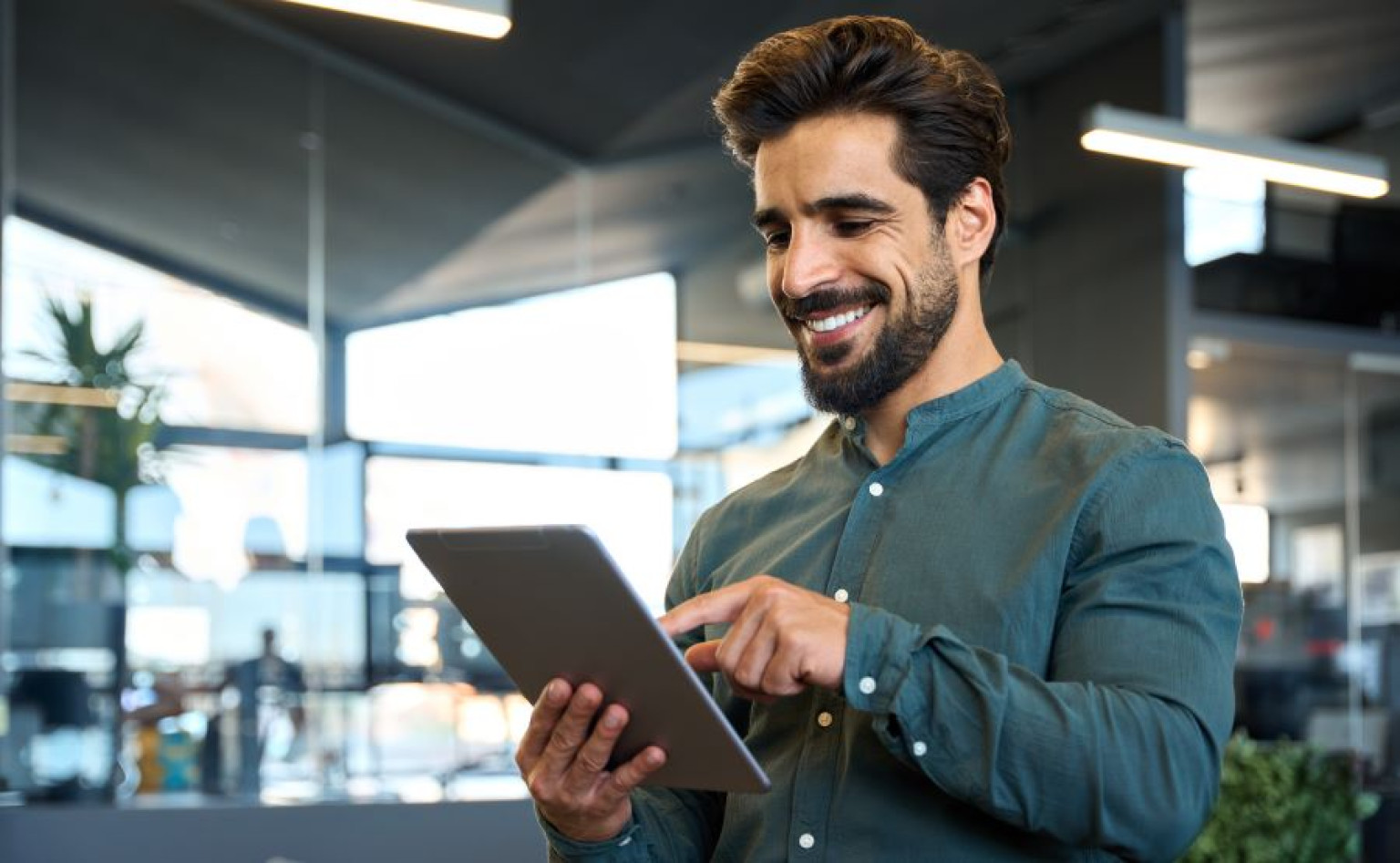 Ein junger Mann mit Bart, gekleidet in ein grünes Hemd, lächelt und bedient ein Tablet in einem modernen Büro. Die Szene vermittelt eine produktive und positive Arbeitsatmosphäre. Als Absolvent des MBA-Programms mit Schwerpunkt Künstliche Intelligenz besitzt er das Wissen, um innovative KI-Lösungen in die Unternehmensstrategie zu integrieren und zukunftsorientierte Karrierewege einzuschlagen.