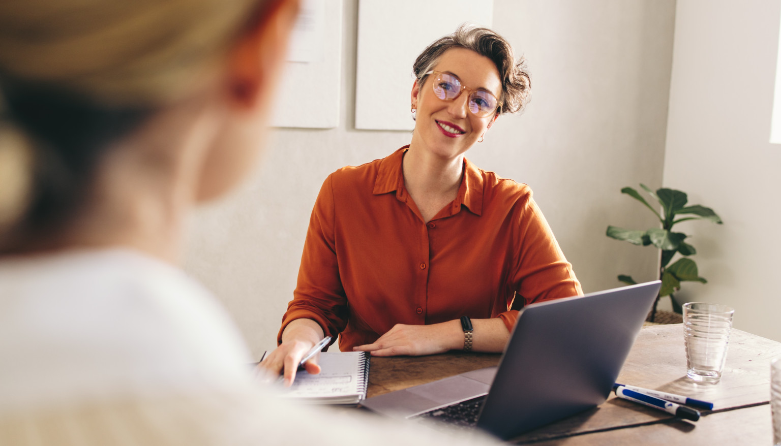 Eine Person sitzt an einem Tisch und führt ein Gespräch mit einer anderen Person, die nicht im Bild zu sehen ist. Die Person im Vordergrund, eine Absolventin des MBA Management – Marketing der Brand University, trägt ein orangefarbenes Hemd und eine Brille und lächelt freundlich.