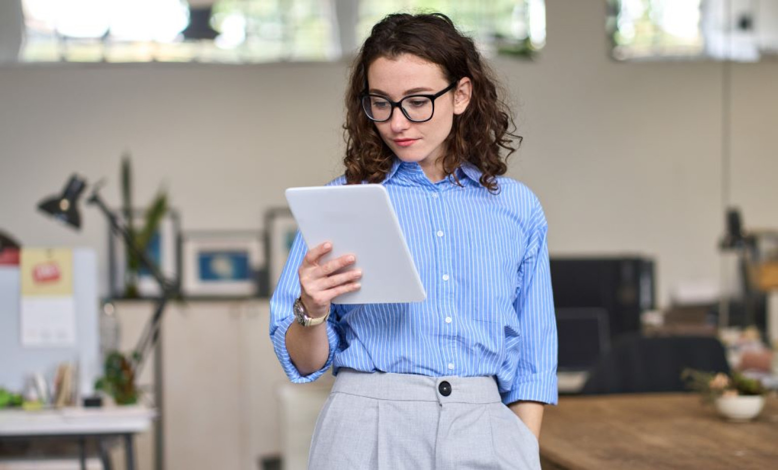 Eine Absolventin des MBA Management – Media & Business Communications steht in einem modernen Büro und schaut auf ein Tablet, das sie in den Händen hält. Sie trägt ein blau-weiß gestreiftes Hemd und eine graue Hose. Im Hintergrund sind Büromöbel und Pflanzen zu sehen, was eine kreative Arbeitsumgebung vermittelt.