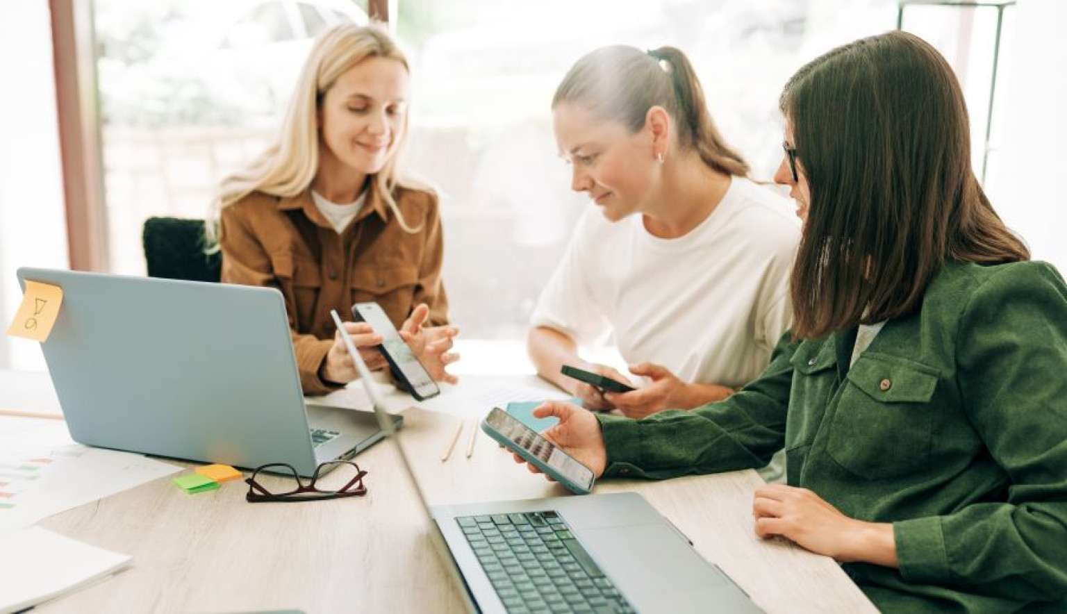 Drei Frauen sitzen an einem Tisch und schauen auf ihre Smartphones, während sie miteinander kommunizieren. Auf dem Tisch liegen ein Laptop, Brillen und farbige Haftnotizen. Im Hintergrund ist Tageslicht sichtbar, das durch große Fenster strömt. Das Bild vermittelt eine kollaborative Atmosphäre. Sie unterhalten sich zum Thema Medienplanung für das nächstes Quartal.