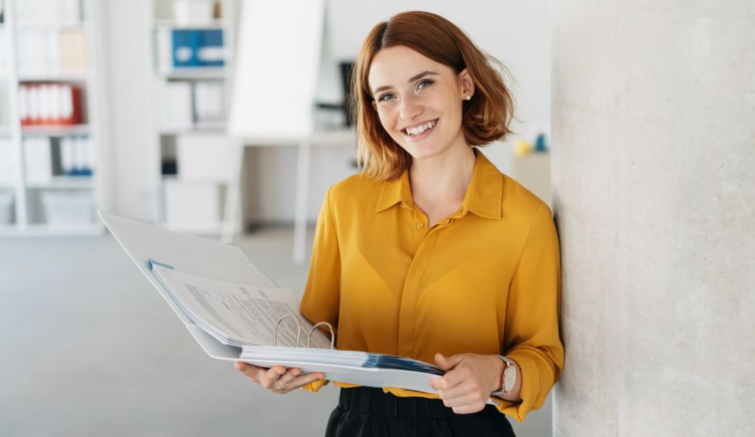 Eine Person steht in einem modernen Büro und hält einen Ordner mit Dokumenten in den Händen. Sie trägt ein gelbes Hemd und lächelt freundlich in die Kamera. Der Hintergrund zeigt Regale mit Akten und eine minimalistische Wand. Das Thema des Bildes bezieht sich auf die Karriereperspektiven im Bereich Nachhaltigkeit im Rahmen eines MBA-Programms, das sich auf nachhaltige Geschäftspraktiken und umweltbewusste Strategien konzentriert.