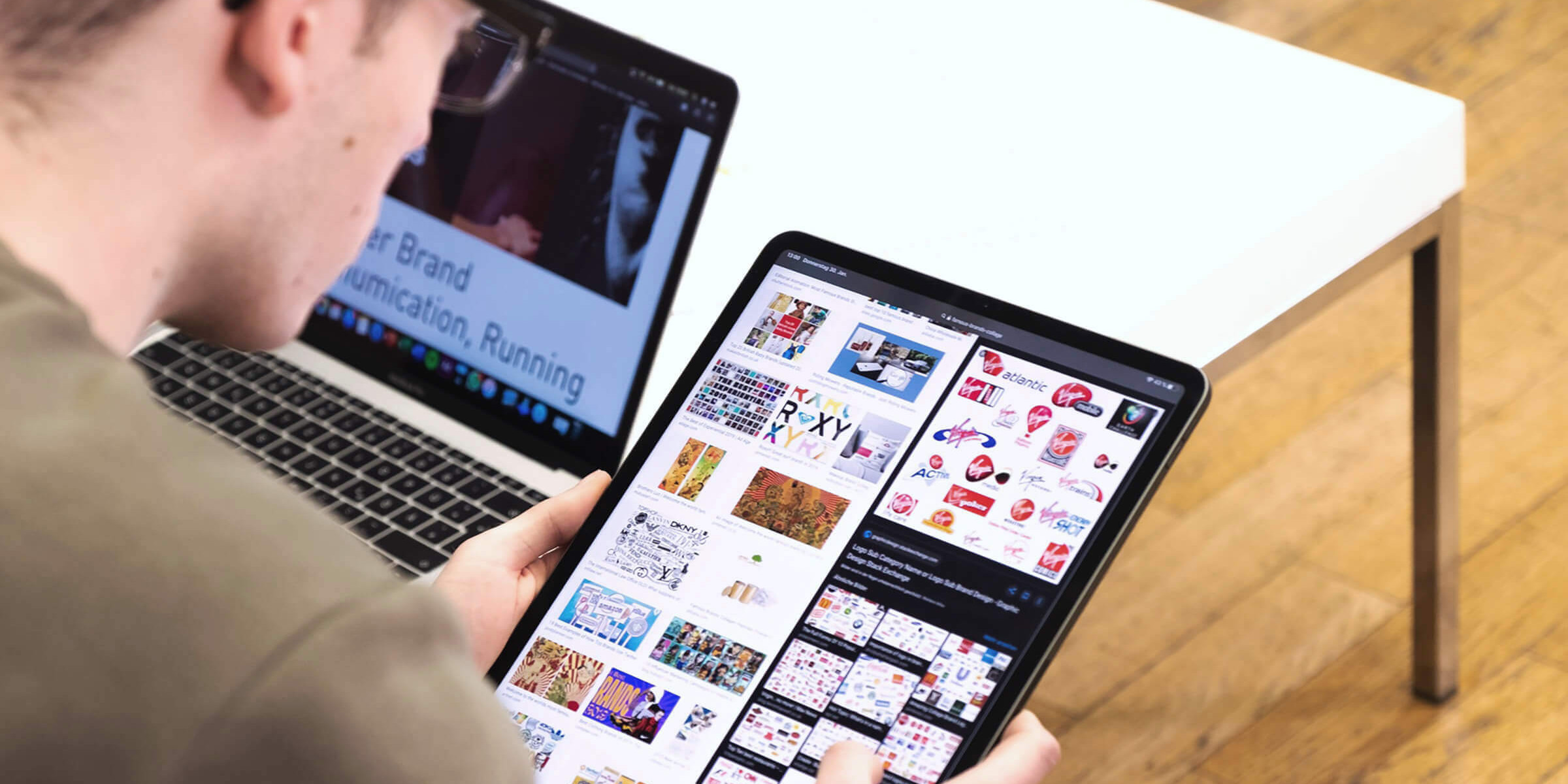 Das Bild zeigt eine Person, die vor einem Laptop sitzt und ein Tablet in den Händen hält. Auf dem Tablet sind verschiedene Bilder und Grafiken zu sehen, möglicherweise im Rahmen eines kreativen Projekts oder einer Bildauswahl. Der Laptop im Hintergrund zeigt eine geöffnete Website oder Präsentation. Die Person scheint sich auf die Auswahl der Inhalte zu konzentrieren. Der Raum hat einen Holzboden und einen schlichten weißen Tisch, was auf eine minimalistische und moderne Arbeitsumgebung hindeutet. Die Szene deutet auf eine digitale Arbeitsweise hin, bei der sowohl Laptop als auch Tablet genutzt werden.