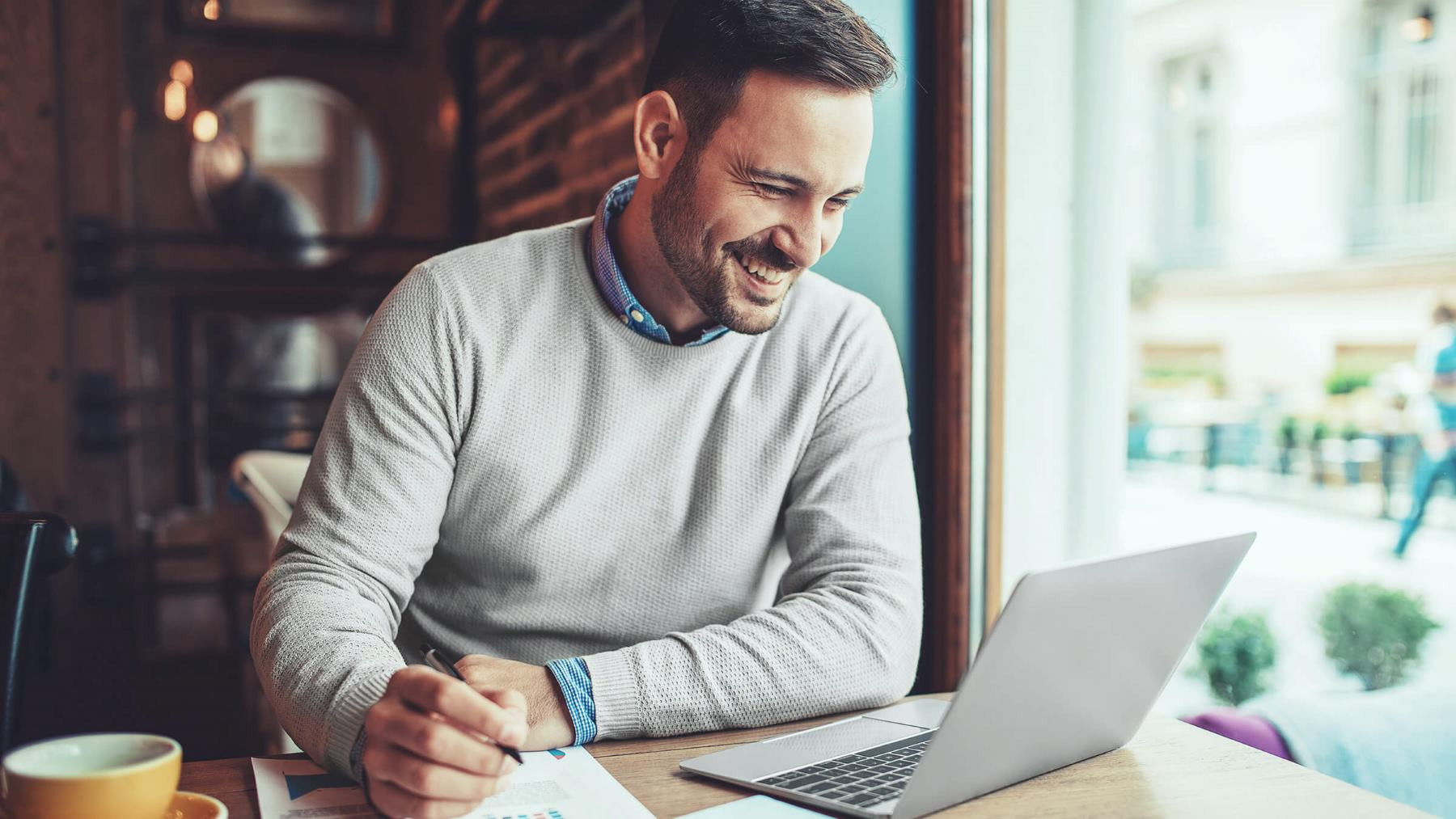 Das Bild zeigt einen lächelnden Mann, der an einem Tisch in einem Café sitzt und an einem Laptop arbeitet. Er trägt einen grauen Pullover über einem Hemd und wirkt konzentriert und gut gelaunt, während er auf den Bildschirm schaut. Neben ihm liegen ein Notizbuch und ein Stift, was darauf hindeutet, dass er möglicherweise Notizen macht oder an einem Projekt arbeitet. Eine Tasse Kaffee steht ebenfalls auf dem Tisch, was die entspannte Atmosphäre unterstreicht. Durch das Fenster ist die Außenseite des Cafés zu sehen, was darauf hinweist, dass der Mann in einer gemütlichen Umgebung arbeitet, möglicherweise im Rahmen eines mobilen oder flexiblen Arbeitsplatzes.