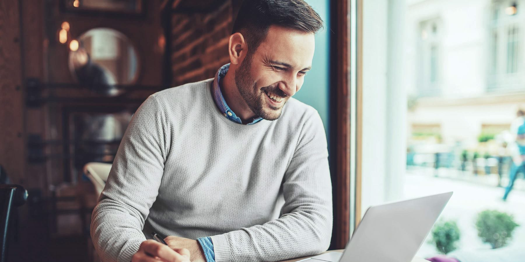 Das Bild zeigt einen lächelnden Mann, der an einem Tisch in einem Café sitzt und an einem Laptop arbeitet. Er trägt einen grauen Pullover über einem Hemd und wirkt konzentriert und gut gelaunt, während er auf den Bildschirm schaut. Neben ihm liegen ein Notizbuch und ein Stift, was darauf hindeutet, dass er möglicherweise Notizen macht oder an einem Projekt arbeitet. Eine Tasse Kaffee steht ebenfalls auf dem Tisch, was die entspannte Atmosphäre unterstreicht. Durch das Fenster ist die Außenseite des Cafés zu sehen, was darauf hinweist, dass der Mann in einer gemütlichen Umgebung arbeitet, möglicherweise im Rahmen eines mobilen oder flexiblen Arbeitsplatzes.