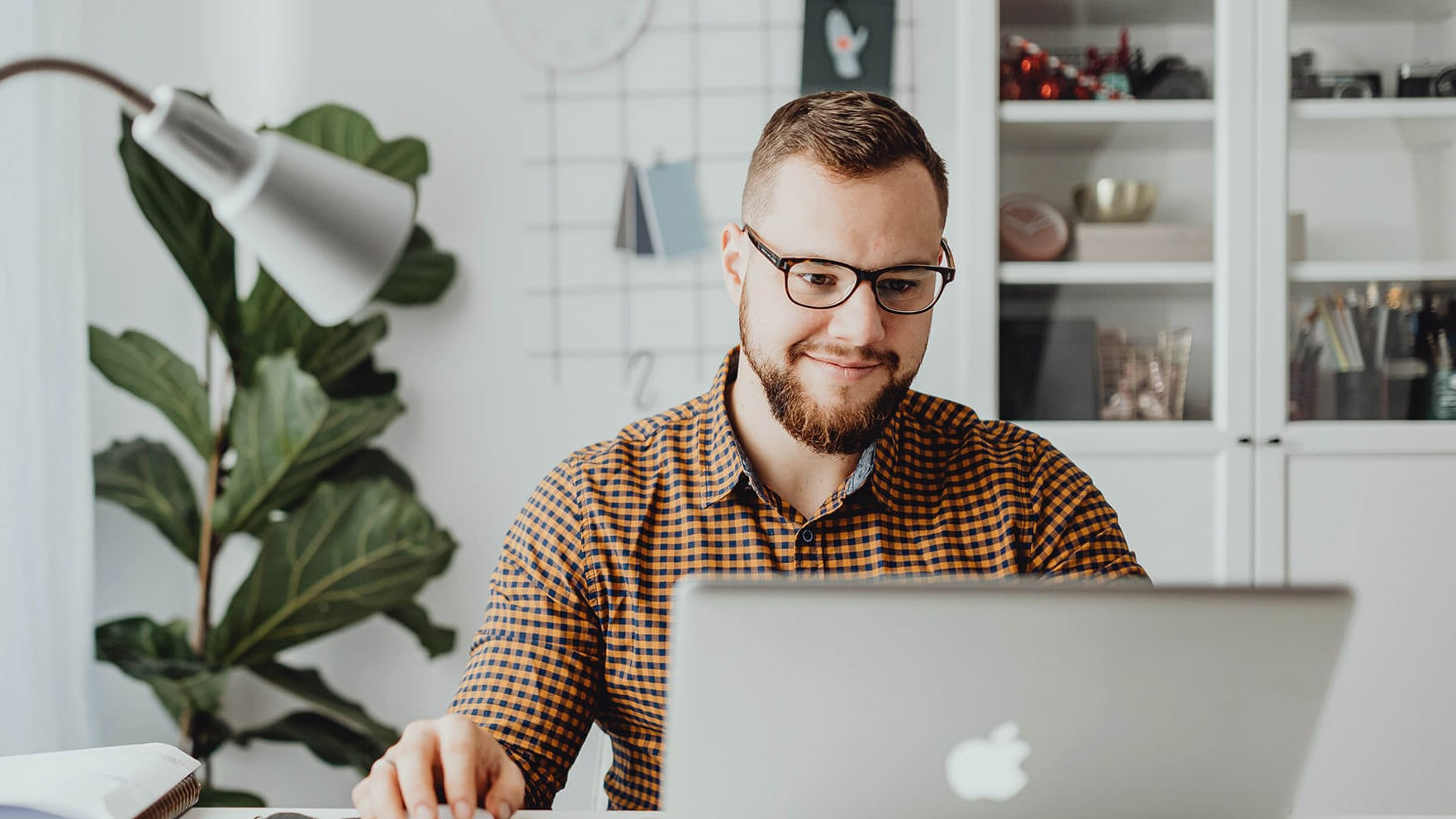 Das Bild zeigt einen Mann mit Brille und Bart, der an einem Schreibtisch vor einem Laptop sitzt und konzentriert arbeitet. Er trägt ein kariertes Hemd und bedient die Computermaus, während er auf den Laptop-Bildschirm schaut. Im Hintergrund sind Regale mit Büchern und Dekorationen zu sehen sowie eine große Pflanze, die dem Raum eine gemütliche und moderne Atmosphäre verleiht. Auf dem Schreibtisch liegen mehrere Gegenstände wie Notizbücher, Stifte und ein Telefon, was auf eine organisierte und produktive Arbeitsumgebung hindeutet. Der Mann wirkt entspannt und fokussiert in seiner Arbeit.