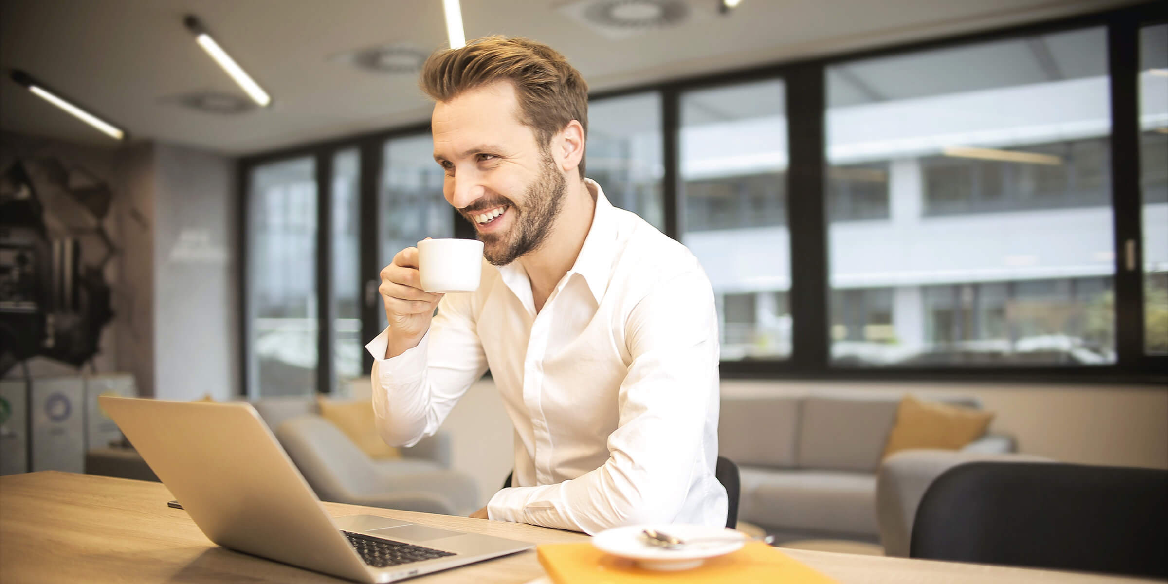 Das Bild zeigt einen lächelnden Mann, der an einem Schreibtisch in einem modernen Büro sitzt und eine Tasse Kaffee trinkt. Er trägt ein weißes Hemd und hat einen Laptop vor sich geöffnet. Auf dem Tisch liegen ein Notizbuch und einige Unterlagen, was auf eine produktive Arbeitssituation hinweist. Im Hintergrund sind große Fenster und ein gemütlicher Sitzbereich mit Sofas zu sehen, was eine offene und angenehme Arbeitsatmosphäre vermittelt. Der Mann wirkt entspannt und zufrieden, möglicherweise genießt er eine kurze Pause während der Arbeit.