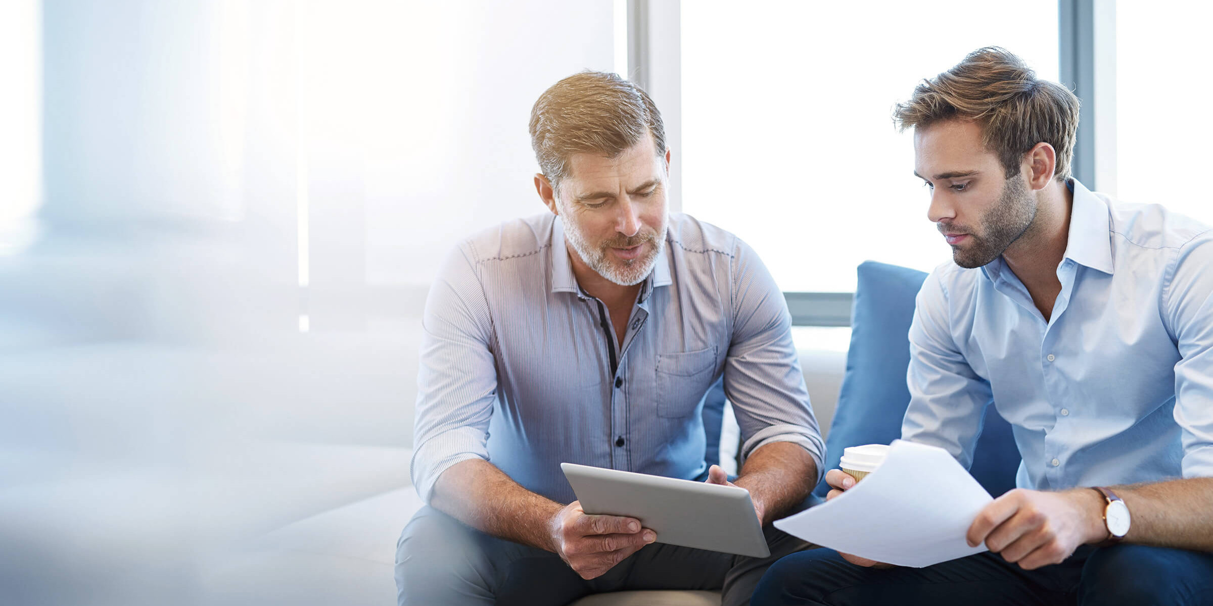 Das Bild zeigt zwei Männer, die gemeinsam auf einem Sofa sitzen und konzentriert an einem Tablet arbeiten. Der Mann links hält das Tablet, während der Mann rechts ein Dokument in der Hand hat. Beide scheinen in ein professionelles Gespräch vertieft zu sein, möglicherweise besprechen sie ein Projekt oder eine Strategie. Sie tragen Hemden, was auf ein Geschäftsumfeld oder ein Meeting hindeutet. Der Hintergrund ist hell und modern gestaltet, und die Atmosphäre wirkt professionell und kollaborativ.