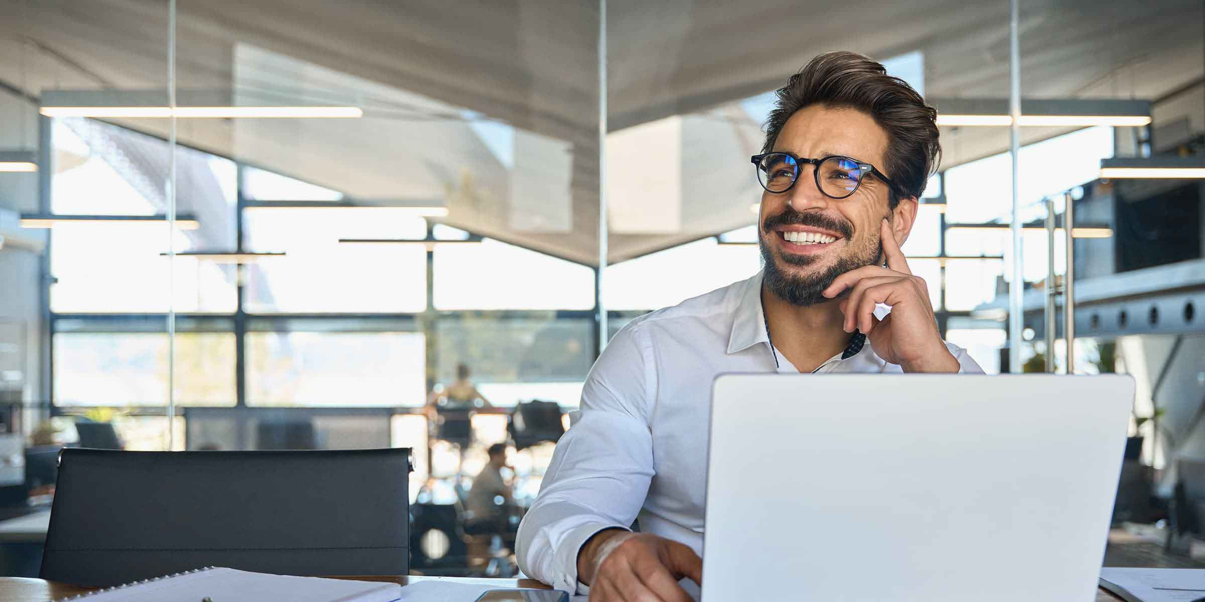 Das Bild zeigt einen lächelnden Mann, der an einem Schreibtisch in einem modernen, gut beleuchteten Büro sitzt. Er trägt ein weißes Hemd und eine Brille und arbeitet an einem Laptop. Neben dem Laptop liegen ein Notizbuch, ein Smartphone und weitere Unterlagen, was auf eine produktive Arbeitssituation hinweist. Der Mann wirkt entspannt und zufrieden, als würde er gerade eine Idee oder einen Gedanken reflektieren. Im Hintergrund ist eine helle, offene Büroumgebung mit großen Fenstern und moderner Einrichtung zu sehen, die eine professionelle und angenehme Arbeitsatmosphäre vermittelt.