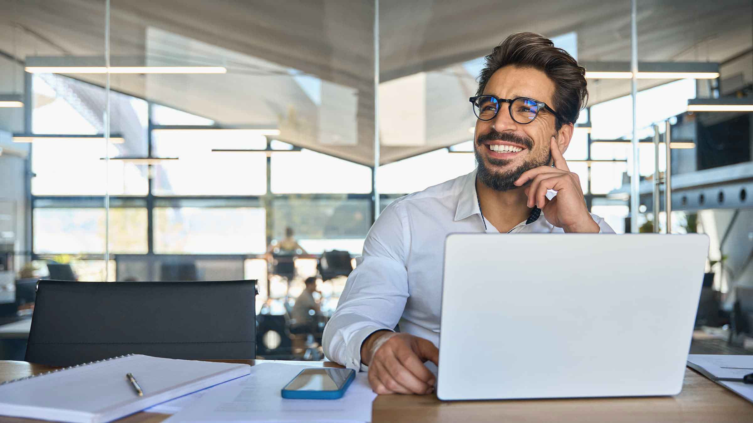 Das Bild zeigt einen lächelnden Mann, der an einem Schreibtisch in einem modernen, gut beleuchteten Büro sitzt. Er trägt ein weißes Hemd und eine Brille und arbeitet an einem Laptop. Neben dem Laptop liegen ein Notizbuch, ein Smartphone und weitere Unterlagen, was auf eine produktive Arbeitssituation hinweist. Der Mann wirkt entspannt und zufrieden, als würde er gerade eine Idee oder einen Gedanken reflektieren. Im Hintergrund ist eine helle, offene Büroumgebung mit großen Fenstern und moderner Einrichtung zu sehen, die eine professionelle und angenehme Arbeitsatmosphäre vermittelt.