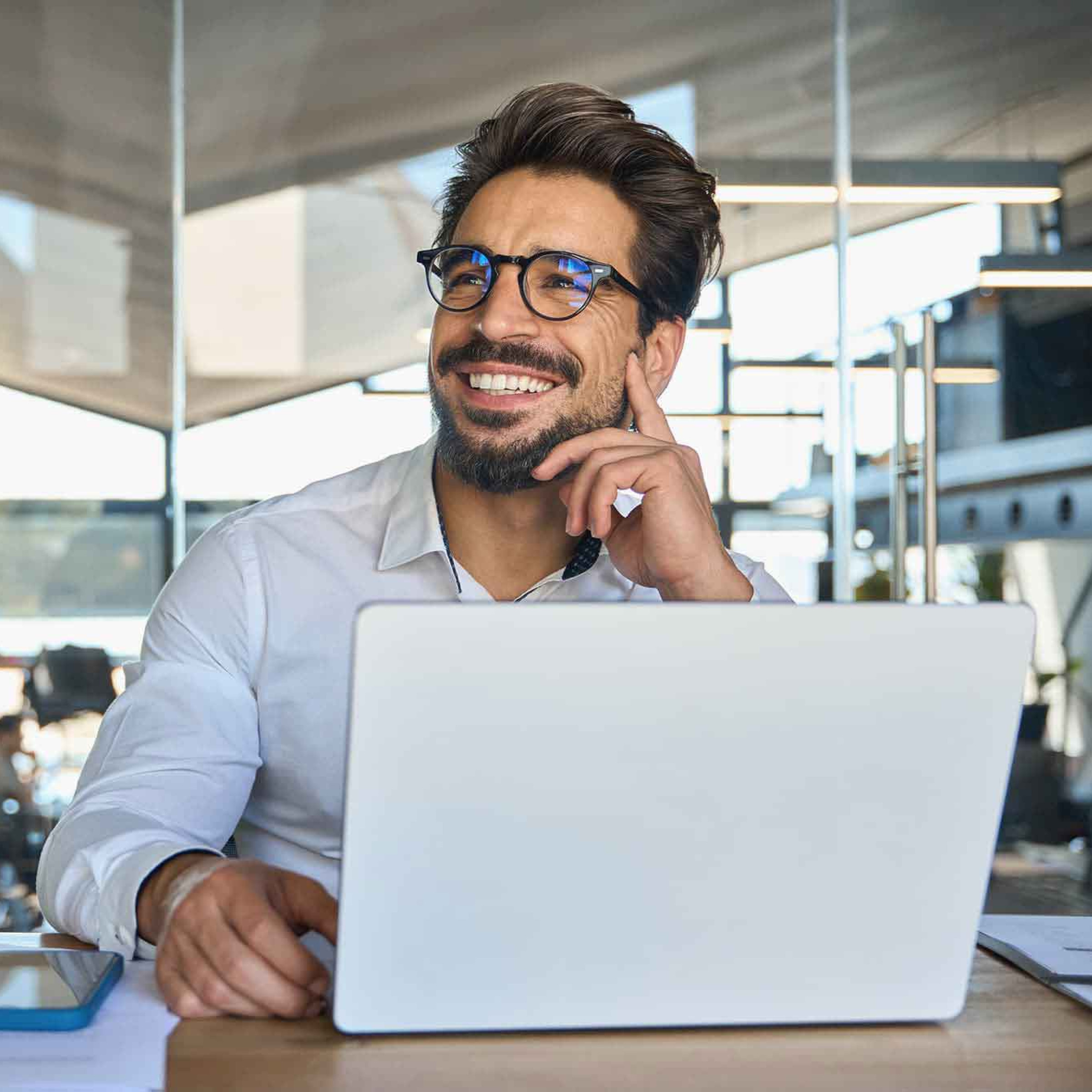 Das Bild zeigt einen lächelnden Mann, der an einem Schreibtisch in einem modernen, gut beleuchteten Büro sitzt. Er trägt ein weißes Hemd und eine Brille und arbeitet an einem Laptop. Neben dem Laptop liegen ein Notizbuch, ein Smartphone und weitere Unterlagen, was auf eine produktive Arbeitssituation hinweist. Der Mann wirkt entspannt und zufrieden, als würde er gerade eine Idee oder einen Gedanken reflektieren. Im Hintergrund ist eine helle, offene Büroumgebung mit großen Fenstern und moderner Einrichtung zu sehen, die eine professionelle und angenehme Arbeitsatmosphäre vermittelt.