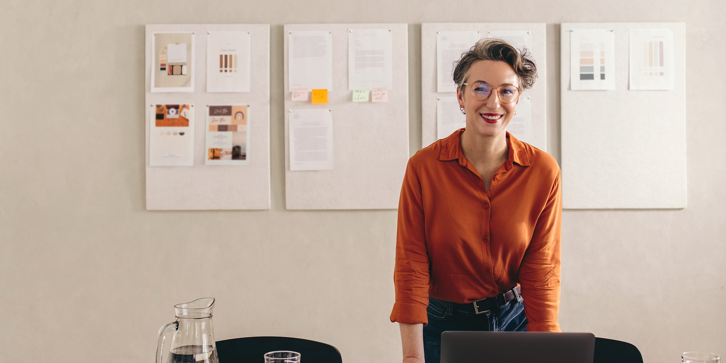 Das Bild zeigt eine Frau mit kurzen Haaren und Brille, die lächelnd an einem Tisch steht. Sie trägt eine orangefarbene Bluse und hat ihre Hände auf dem Tisch abgestützt, während sie in die Kamera schaut. Vor ihr steht ein Laptop, und auf dem Tisch befinden sich zudem eine Karaffe mit Wasser, ein Glas und einige Notizbücher, was auf eine kreative oder produktive Arbeitssituation hindeutet. Im Hintergrund hängen mehrere Dokumente und Notizen an der Wand, die möglicherweise zu einem Projekt oder einer Präsentation gehören. Die Atmosphäre wirkt offen und kreativ, und die Frau scheint mitten in einer professionellen Arbeitsumgebung zu sein.