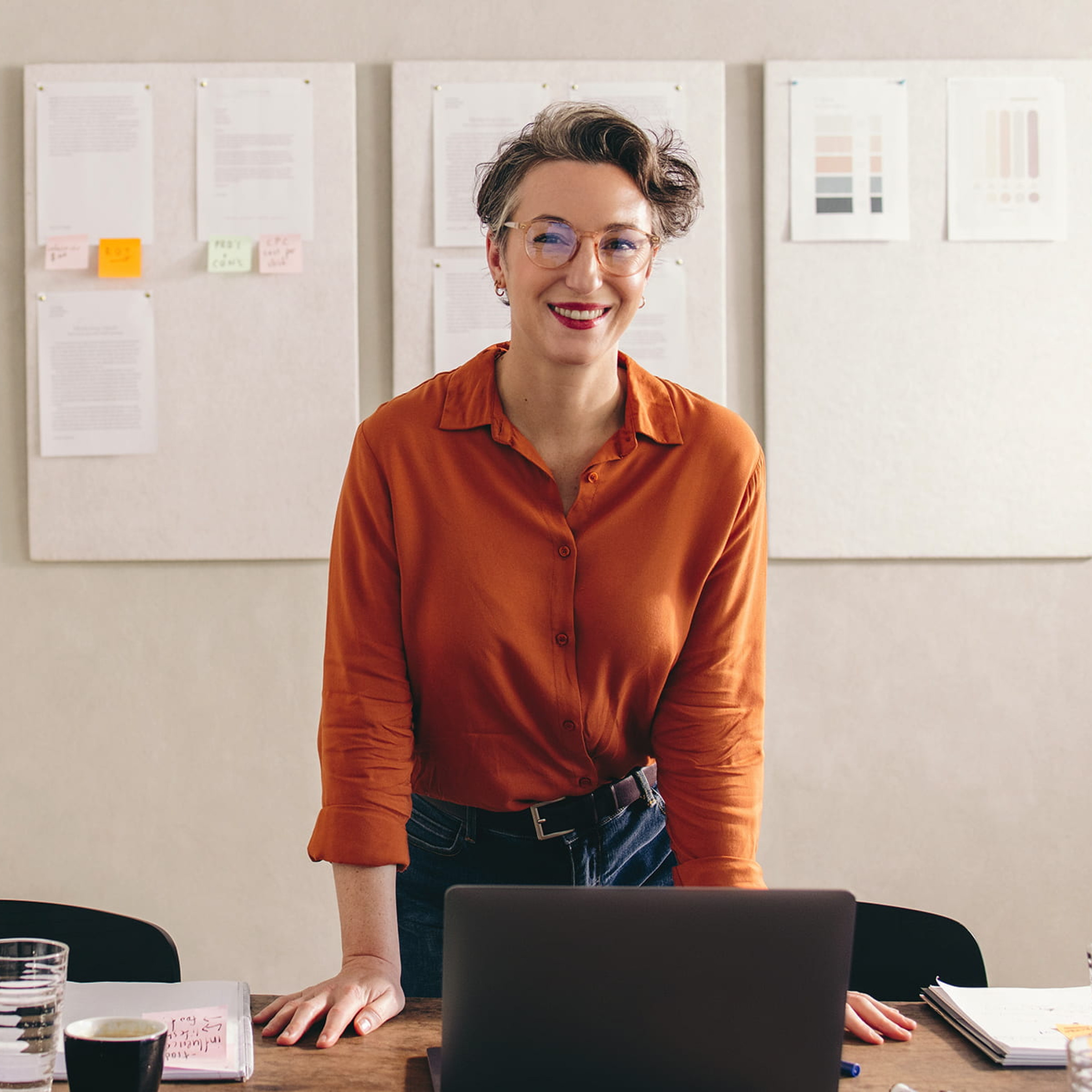 Das Bild zeigt eine Frau mit kurzen Haaren und Brille, die lächelnd an einem Tisch steht. Sie trägt eine orangefarbene Bluse und hat ihre Hände auf dem Tisch abgestützt, während sie in die Kamera schaut. Vor ihr steht ein Laptop, und auf dem Tisch befinden sich zudem eine Karaffe mit Wasser, ein Glas und einige Notizbücher, was auf eine kreative oder produktive Arbeitssituation hindeutet. Im Hintergrund hängen mehrere Dokumente und Notizen an der Wand, die möglicherweise zu einem Projekt oder einer Präsentation gehören. Die Atmosphäre wirkt offen und kreativ, und die Frau scheint mitten in einer professionellen Arbeitsumgebung zu sein.