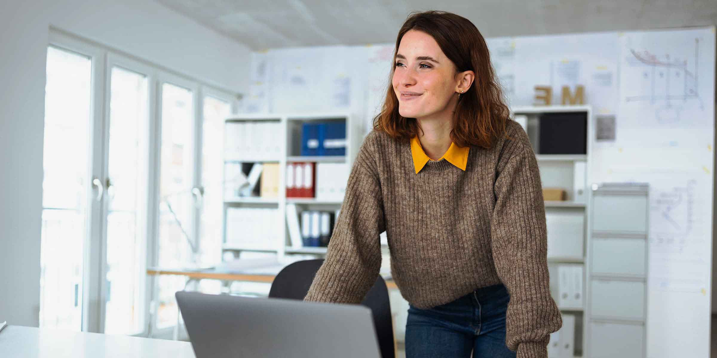 Das Bild zeigt eine junge Frau, die sich lächelnd über einen Tisch lehnt und auf einen Laptop schaut. Sie trägt einen grob gestrickten Pullover in Braun und eine Bluse mit einem gelben Kragen, was ihr Outfit leger und stilvoll wirken lässt. Die Frau steht in einem hellen und gut organisierten Büro mit Regalen, Akten und Plänen an den Wänden im Hintergrund. Der Raum wirkt modern und professionell, und das natürliche Licht, das durch die großen Fenster hereinfällt, verstärkt die freundliche Atmosphäre. Die Frau wirkt konzentriert und zufrieden, als sei sie in ein kreatives oder produktives Projekt vertieft.