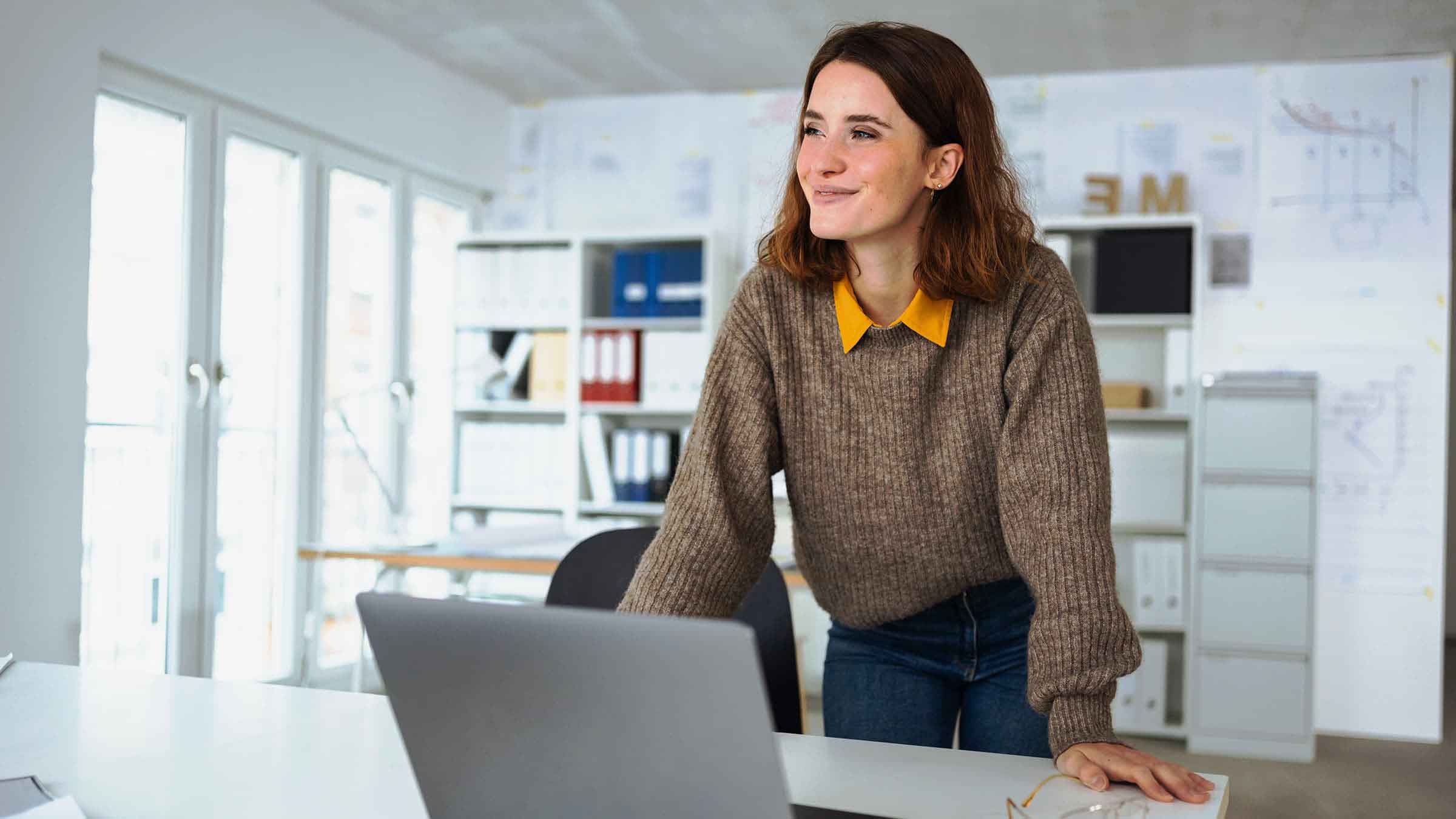 Das Bild zeigt eine junge Frau, die sich lächelnd über einen Tisch lehnt und auf einen Laptop schaut. Sie trägt einen grob gestrickten Pullover in Braun und eine Bluse mit einem gelben Kragen, was ihr Outfit leger und stilvoll wirken lässt. Die Frau steht in einem hellen und gut organisierten Büro mit Regalen, Akten und Plänen an den Wänden im Hintergrund. Der Raum wirkt modern und professionell, und das natürliche Licht, das durch die großen Fenster hereinfällt, verstärkt die freundliche Atmosphäre. Die Frau wirkt konzentriert und zufrieden, als sei sie in ein kreatives oder produktives Projekt vertieft.