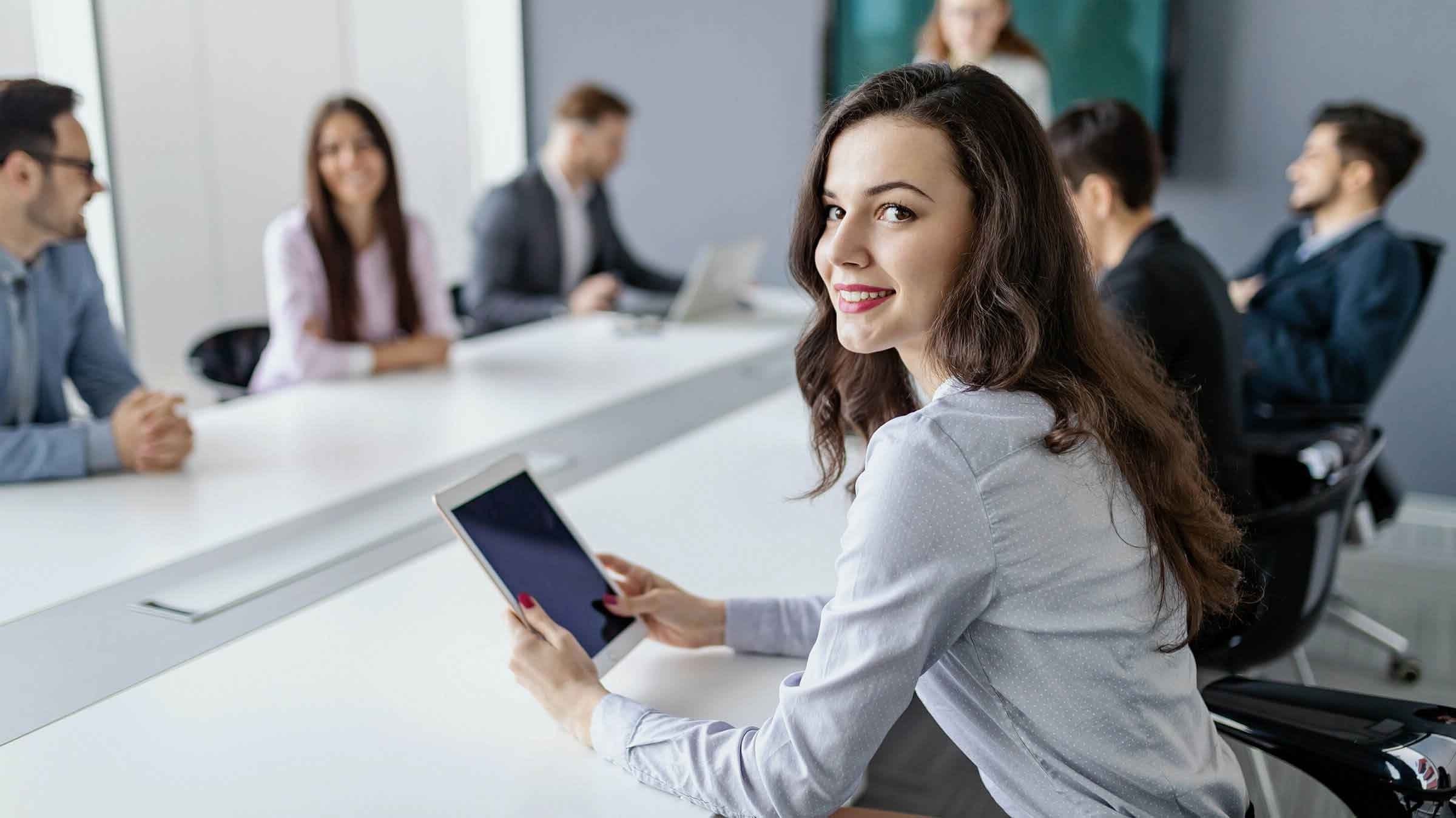 Das Bild zeigt eine junge Frau mit langen, dunklen Haaren, die in einem Konferenzraum an einem Tisch sitzt und ein Tablet in den Händen hält. Sie schaut über die Schulter lächelnd in die Kamera. Um sie herum sitzen mehrere weitere Personen, die in ein Meeting oder eine Diskussion vertieft sind. Die Umgebung wirkt professionell, mit modernen Möbeln und einem großen Besprechungstisch. Die Teilnehmer scheinen in einem formellen Geschäftsumfeld zu arbeiten, und die Atmosphäre vermittelt eine produktive und kollaborative Stimmung. Die Frau wirkt engagiert und freundlich, als ob sie an der Besprechung aktiv teilnimmt.