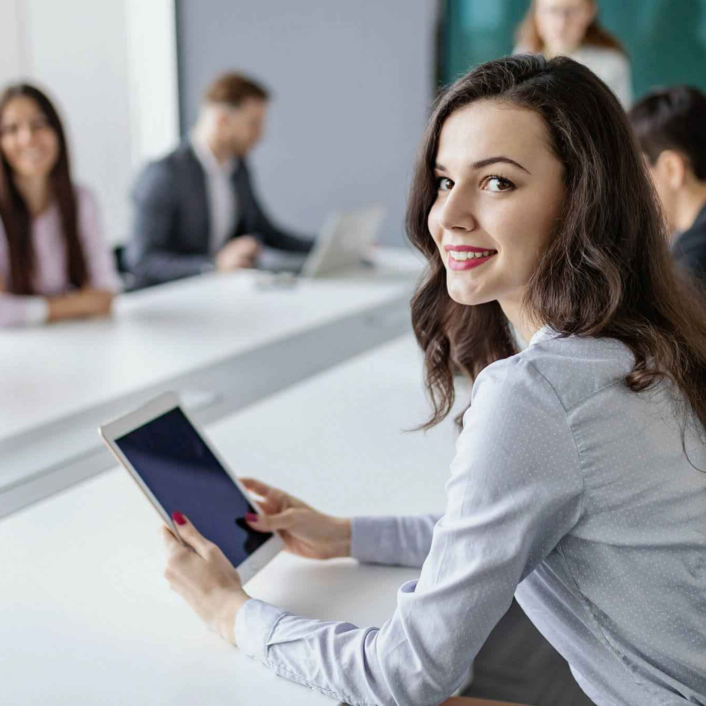 Das Bild zeigt eine junge Frau mit langen, dunklen Haaren, die in einem Konferenzraum an einem Tisch sitzt und ein Tablet in den Händen hält. Sie schaut über die Schulter lächelnd in die Kamera. Um sie herum sitzen mehrere weitere Personen, die in ein Meeting oder eine Diskussion vertieft sind. Die Umgebung wirkt professionell, mit modernen Möbeln und einem großen Besprechungstisch. Die Teilnehmer scheinen in einem formellen Geschäftsumfeld zu arbeiten, und die Atmosphäre vermittelt eine produktive und kollaborative Stimmung. Die Frau wirkt engagiert und freundlich, als ob sie an der Besprechung aktiv teilnimmt.