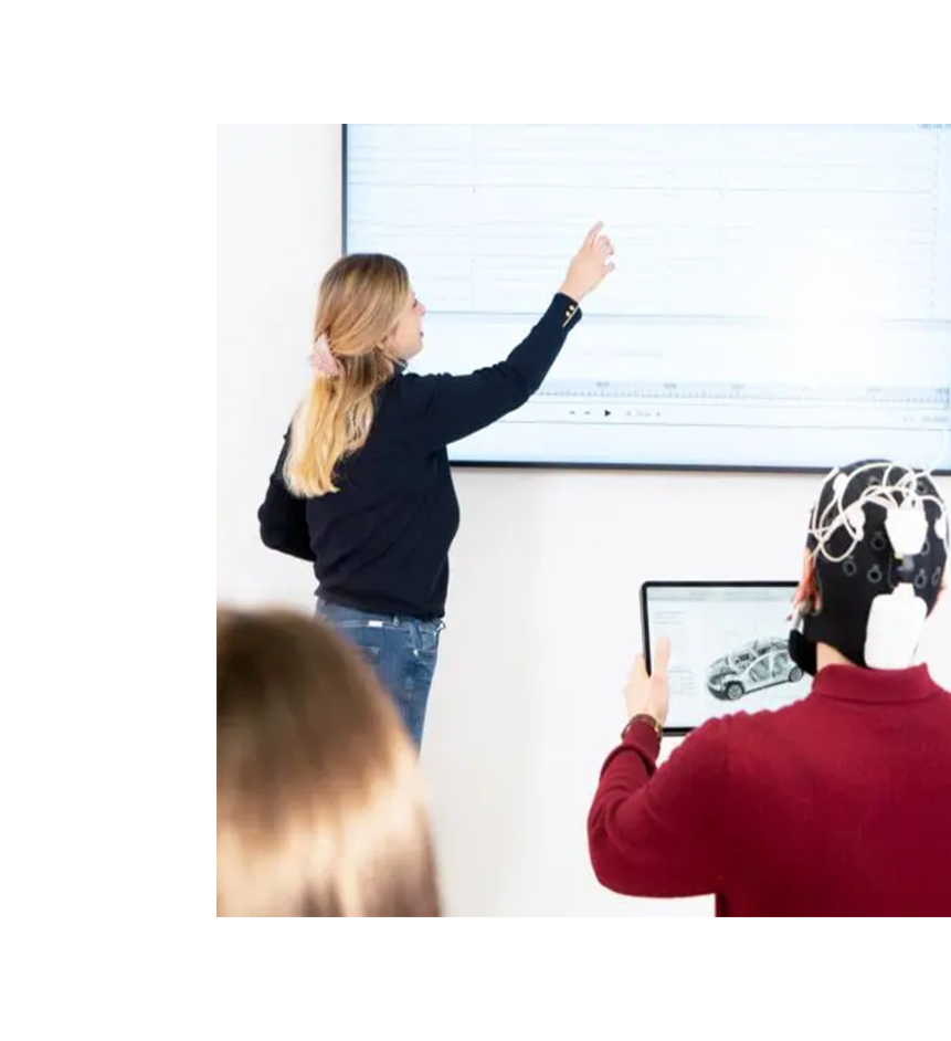 Eine Frau steht vor einem großen Bildschirm und zeigt auf ein Diagramm, während ein Student mit EEG-Kopfbedeckung ein Tablet hält. Die Szene deutet auf eine wissenschaftliche Untersuchung hin, die moderne Technologie und neuropsychologische Methoden einsetzt. Diese innovative Forschungsumgebung spiegelt die praxisorientierten Lernmöglichkeiten an der Brand University wider.