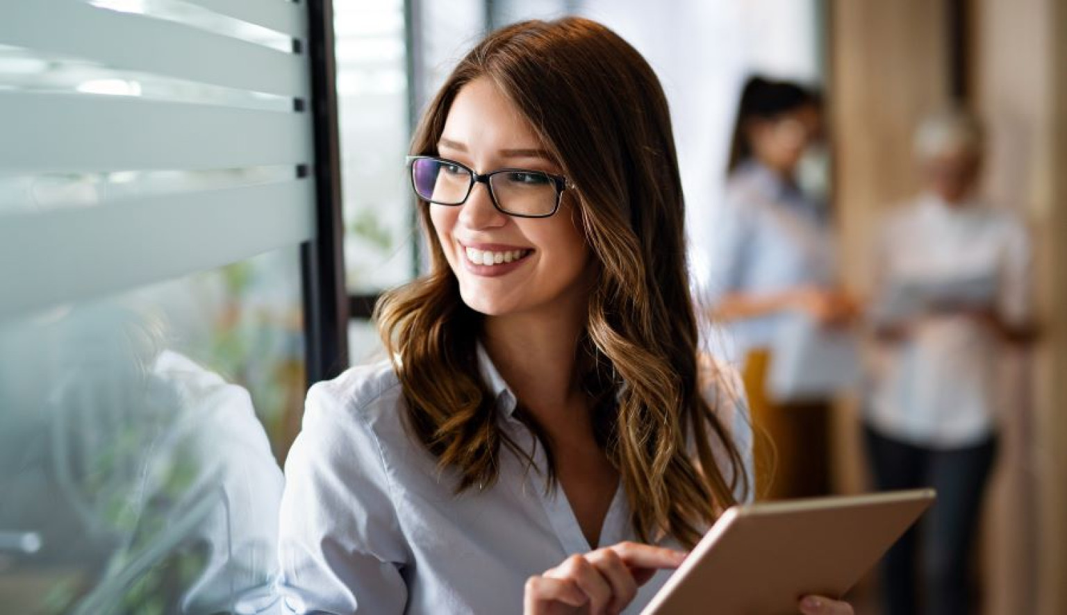 Eine junge Frau mit Brille und Tablet in den Händen lächelt, während sie in einem modernen Büro steht. Im Hintergrund sind verschwommen weitere Personen zu sehen, was auf eine Team- oder Arbeitsumgebung hinweist. Als Absolventin des Fernstudiengangs MBA Digital Business Management bringt sie das Wissen und die Fähigkeiten mit, um digitale Geschäftsstrategien erfolgreich umzusetzen.